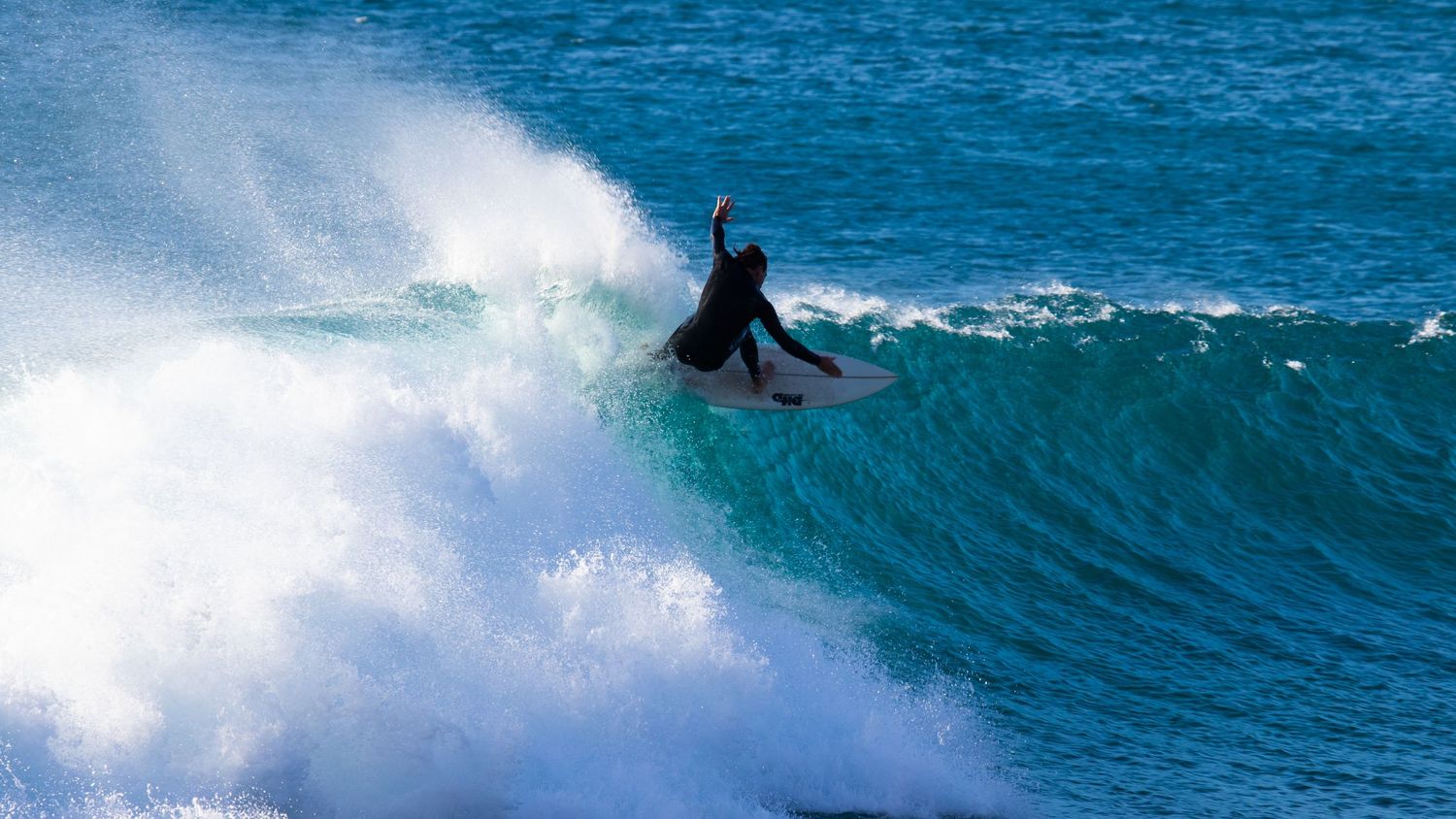 Surfer in black wetsuit carving a sharp turn on a turquoise wave with white foam splashing.