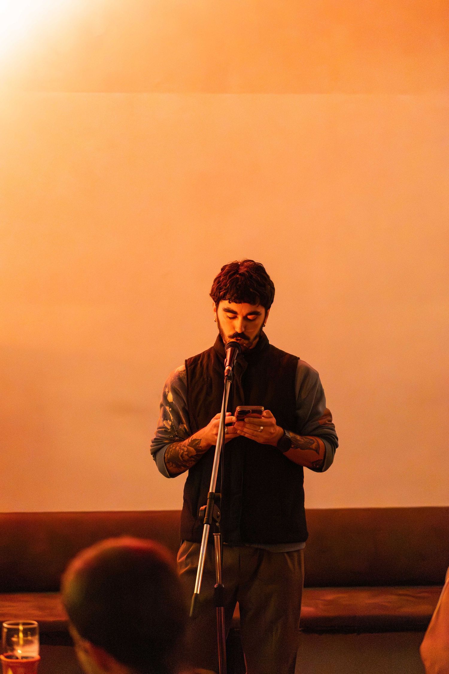 A bearded man reads from his phone at a microphone stand on stage in warm orange lighting.