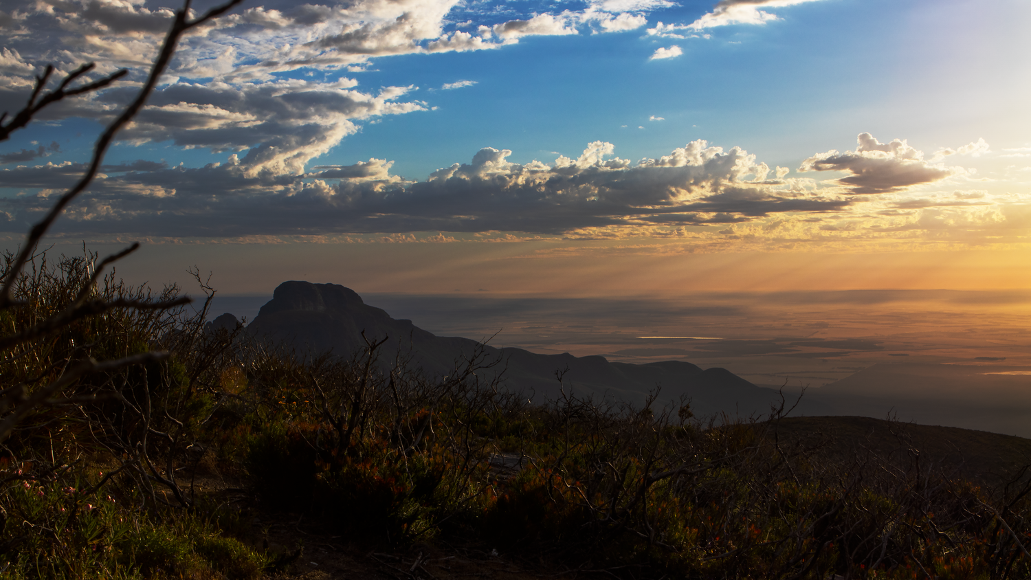 Panoramic mountain sunset view with isolated mesa, golden clouds, and vast plains stretching to the horizon.