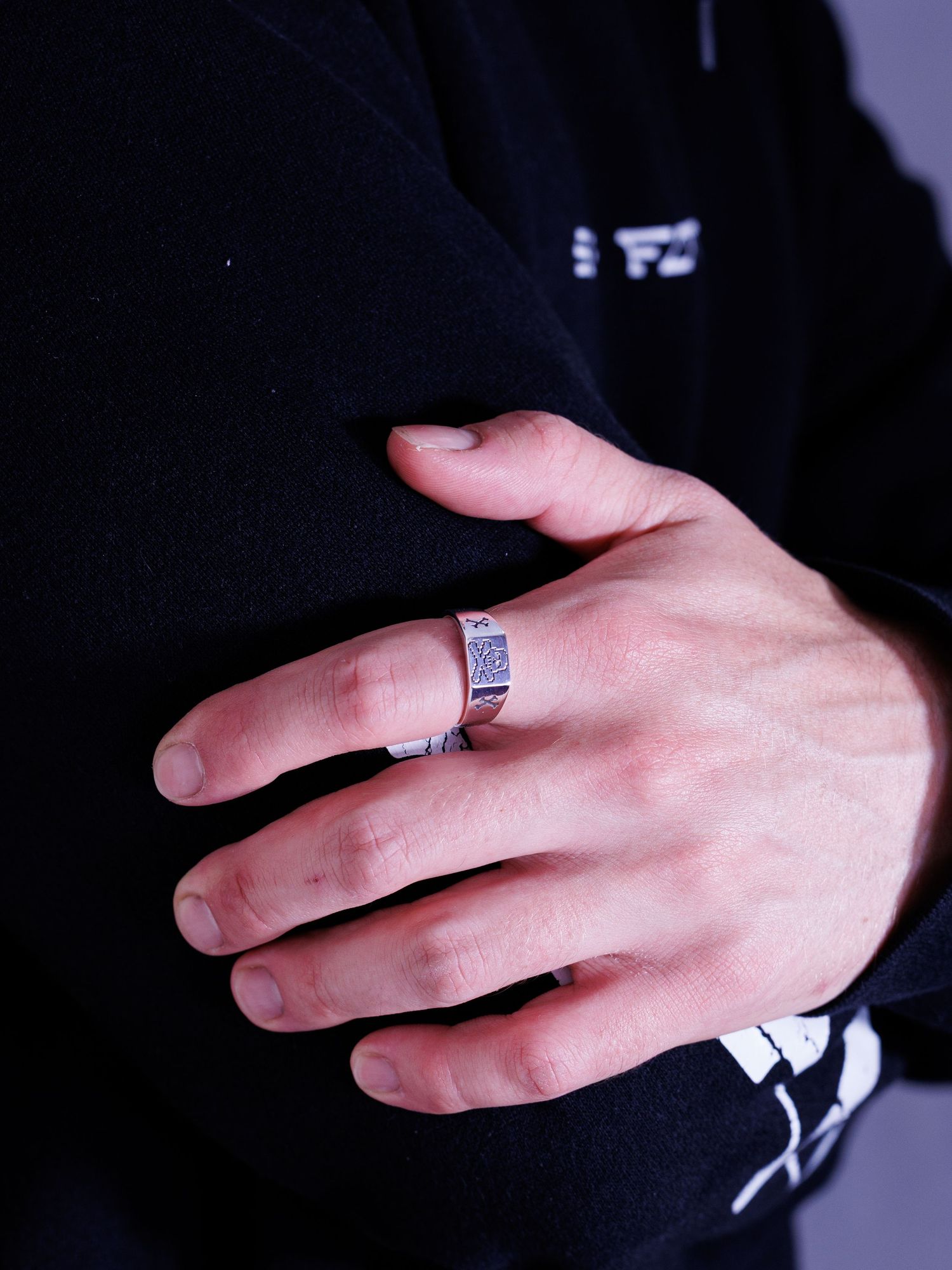 Hand wearing a detailed silver signet ring with engraved design against a dark black hoodie background.