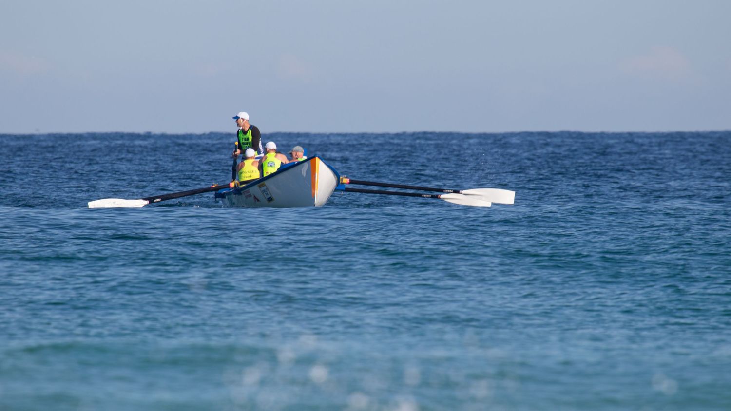 Two surf lifesavers rowing a wooden outrigger canoe on calm blue ocean.