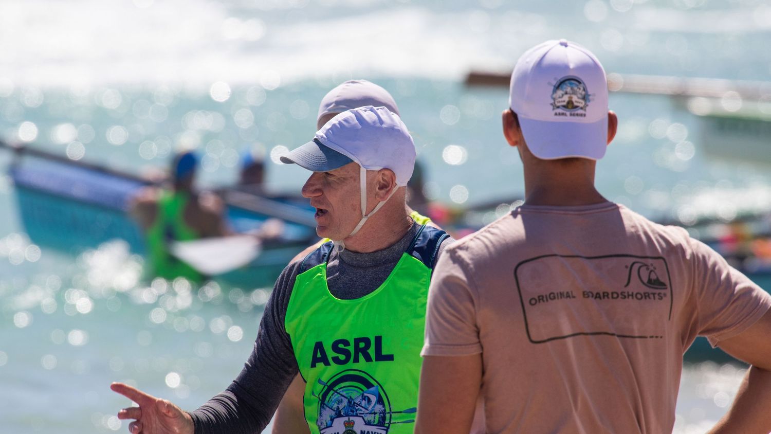 Two surf lifesaving officials in ASRL vests having a discussion near colorful boats on the beach.