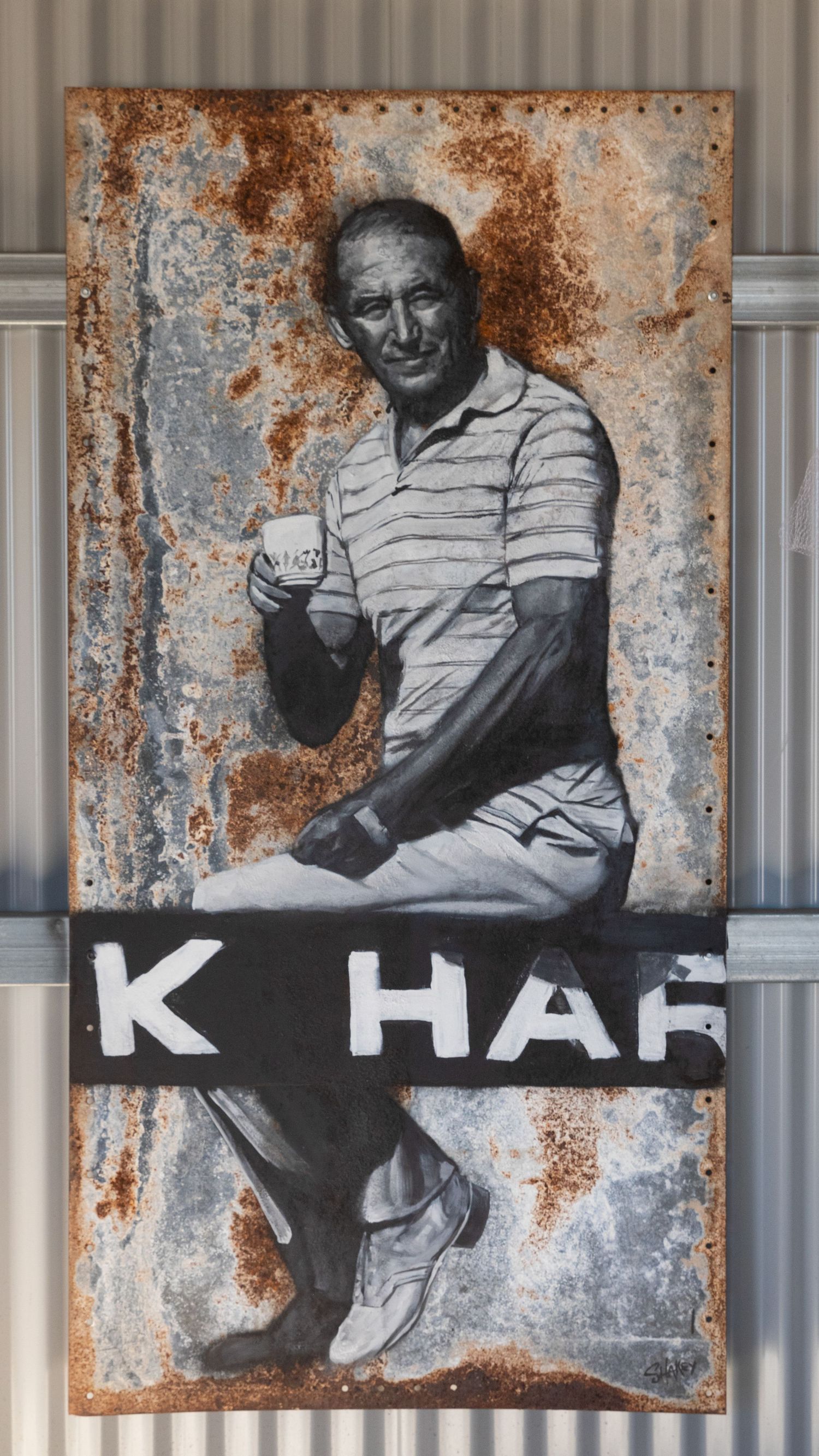 Black and white portrait artwork painted on rusty corrugated iron wall of a man holding a cup.