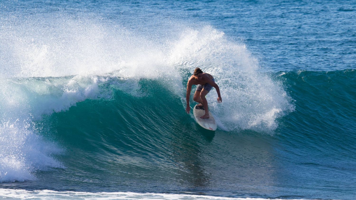 A surfer carves through a large turquoise wave, performing an aggressive turn with water spraying around.