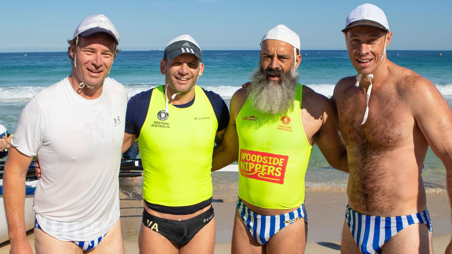 Four smiling surf lifesavers pose on a sunny beach wearing swimwear and white caps at an Australian surf carnival.