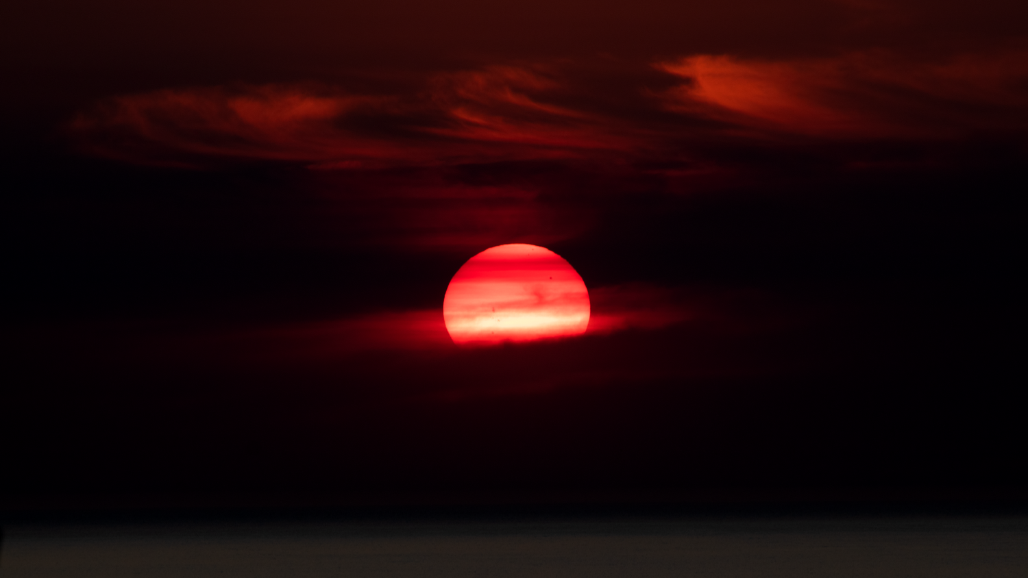 A dramatic red sun sets over dark ocean waters, glowing intensely through moody storm clouds at dusk.