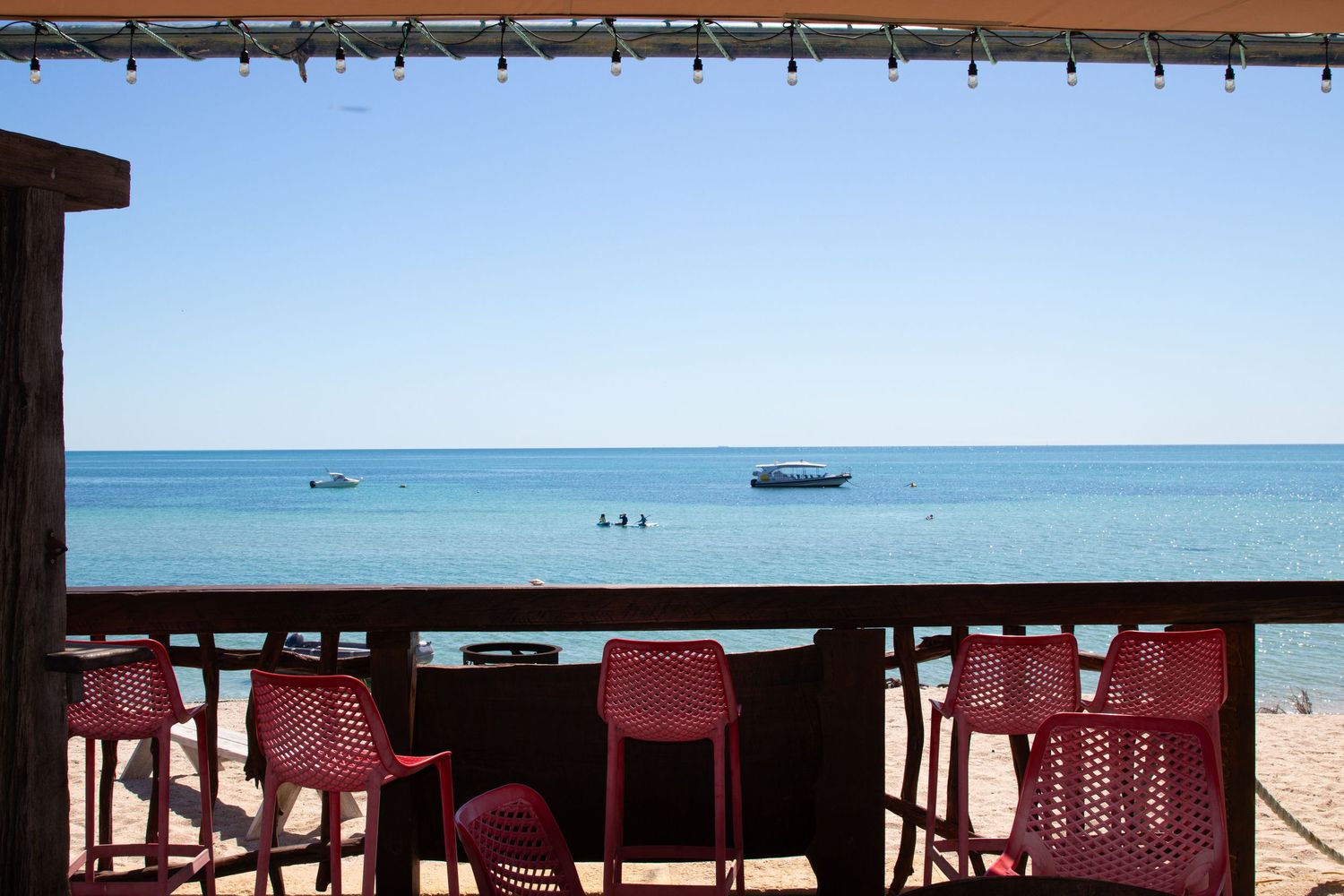 Red bar stools on beachfront deck overlooking calm turquoise ocean with boats anchored on sunny clear day.