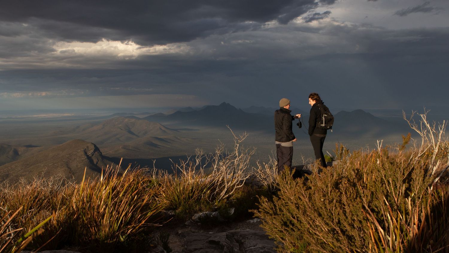 Two hikers conversing on mountain summit with sweeping valley views under stormy clouds.