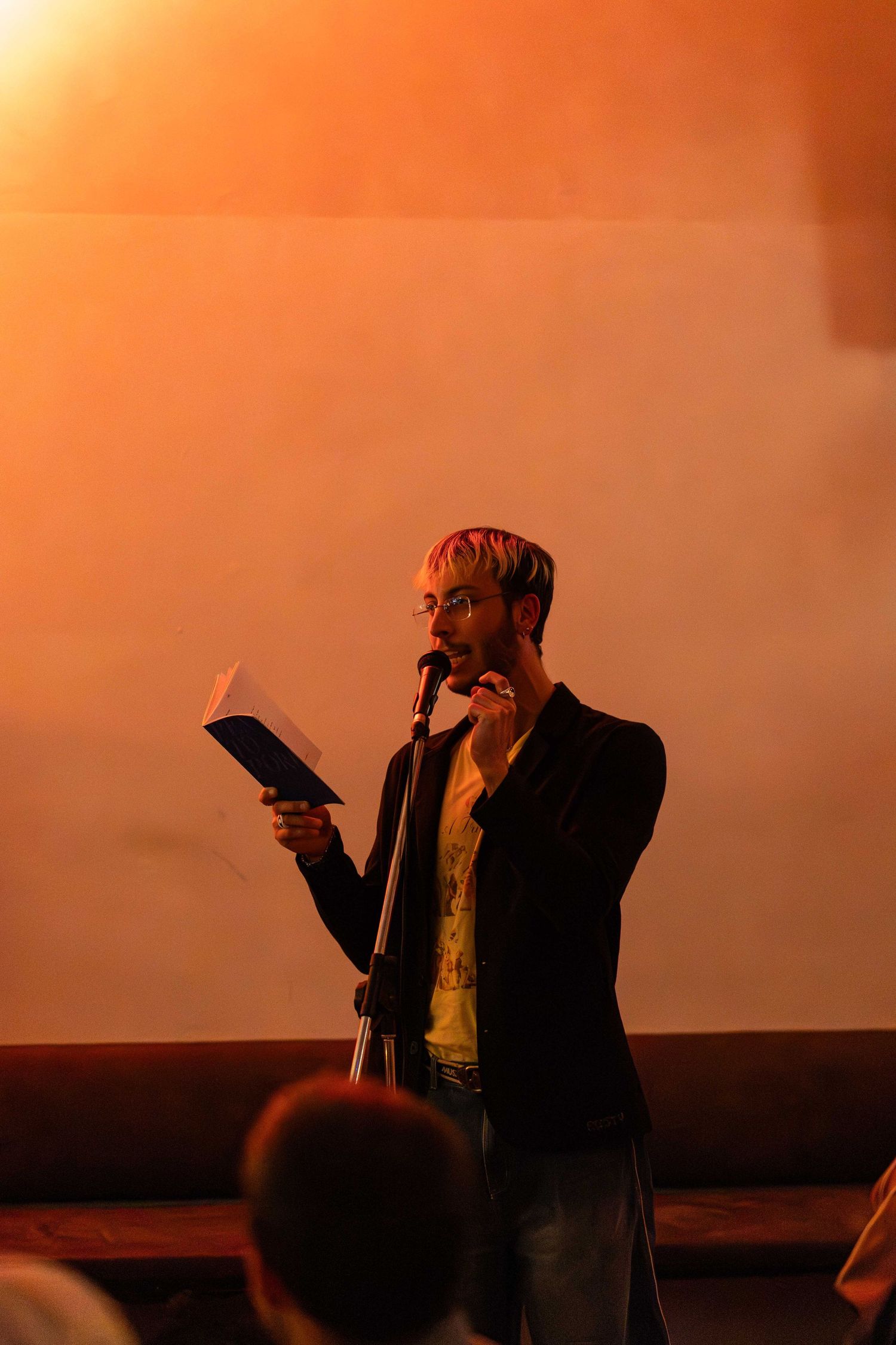 A man reads from a book at a microphone stand on stage, bathed in warm orange lighting before an audience.
