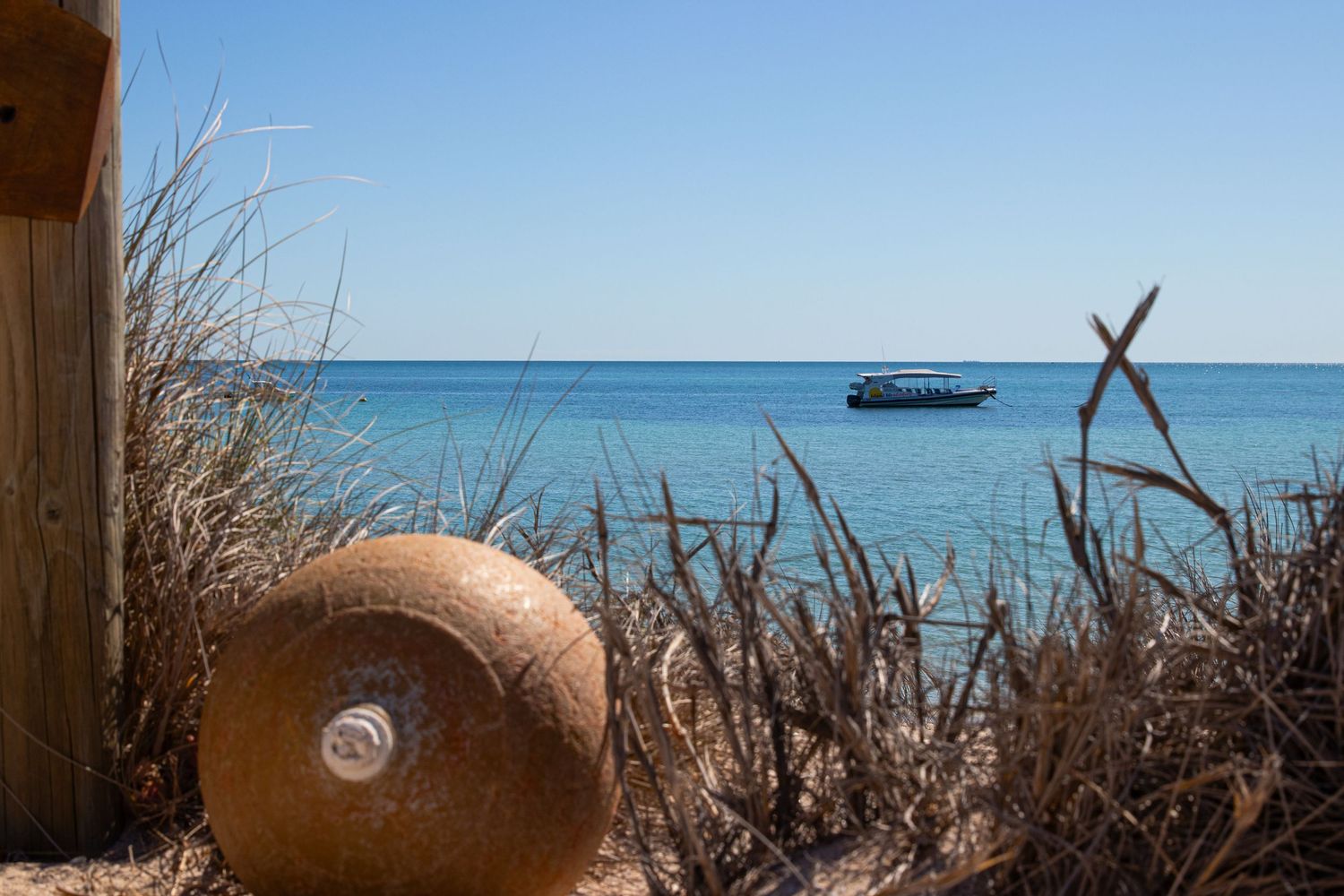 Rustic wooden post and coastal dune grasses frame a turquoise sea with a boat anchored offshore.