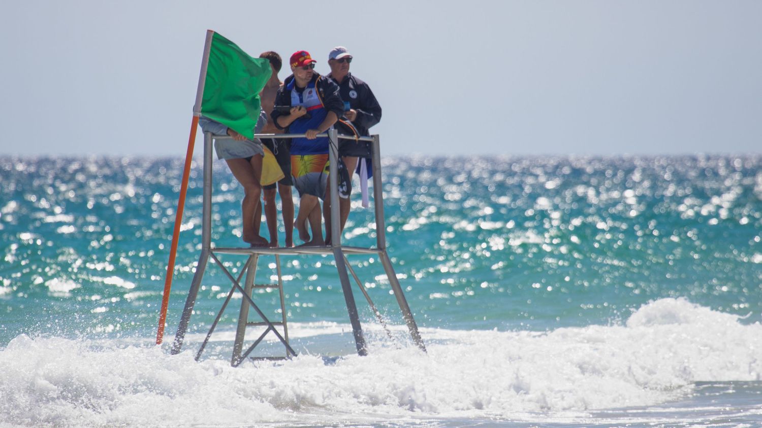 Surf lifesaving officials standing on elevated tower with green flag overlooking choppy ocean waves.