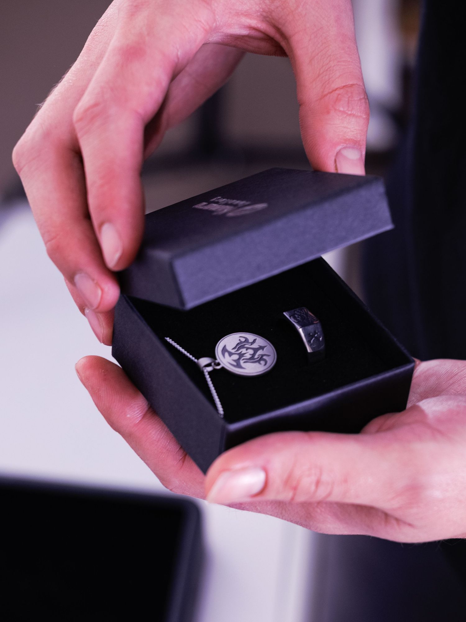 Hands holding an open black jewelry box with a silver engraved pendant necklace and ring inside.