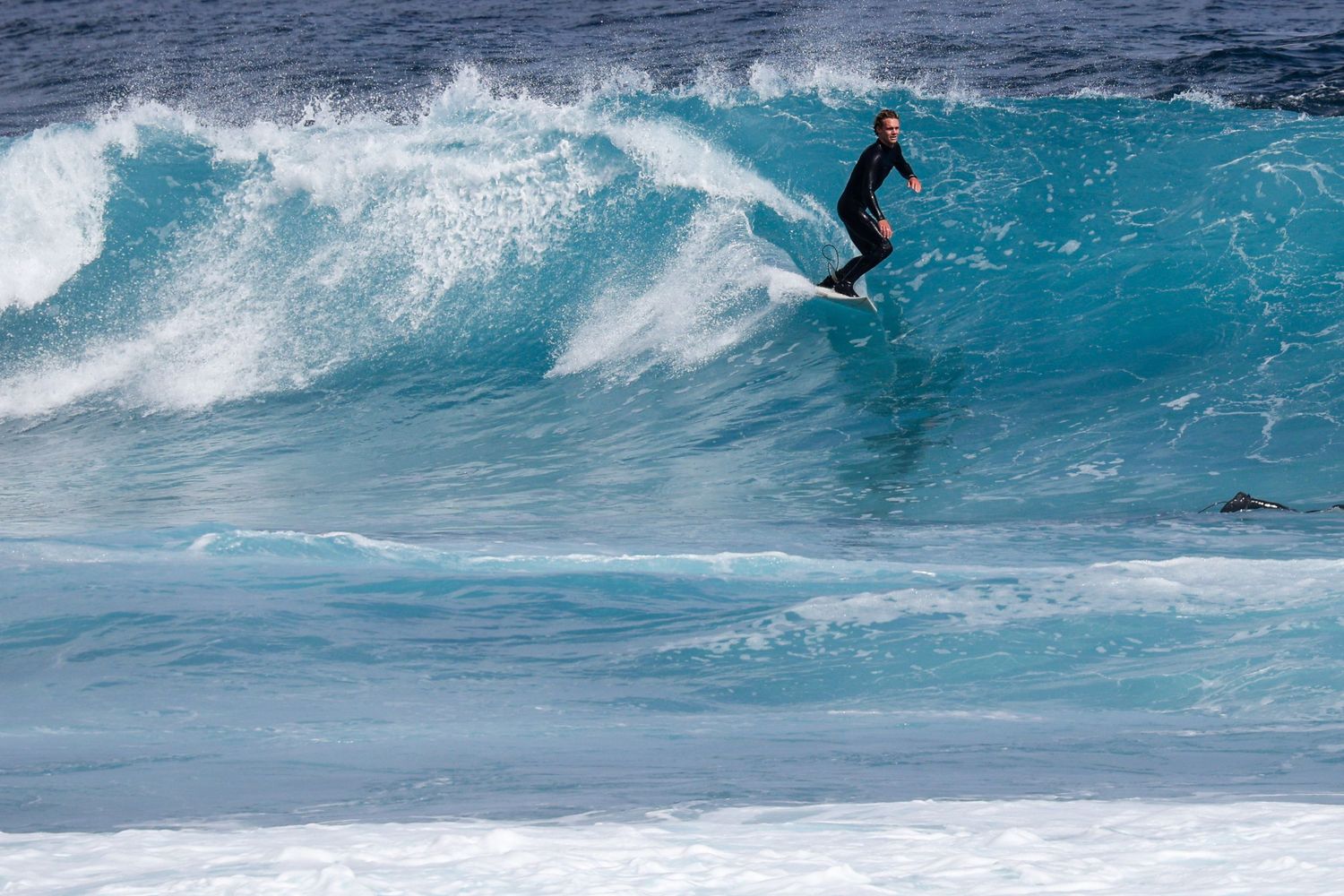 Surfer in a wetsuit riding a large turquoise wave on a clear day.
