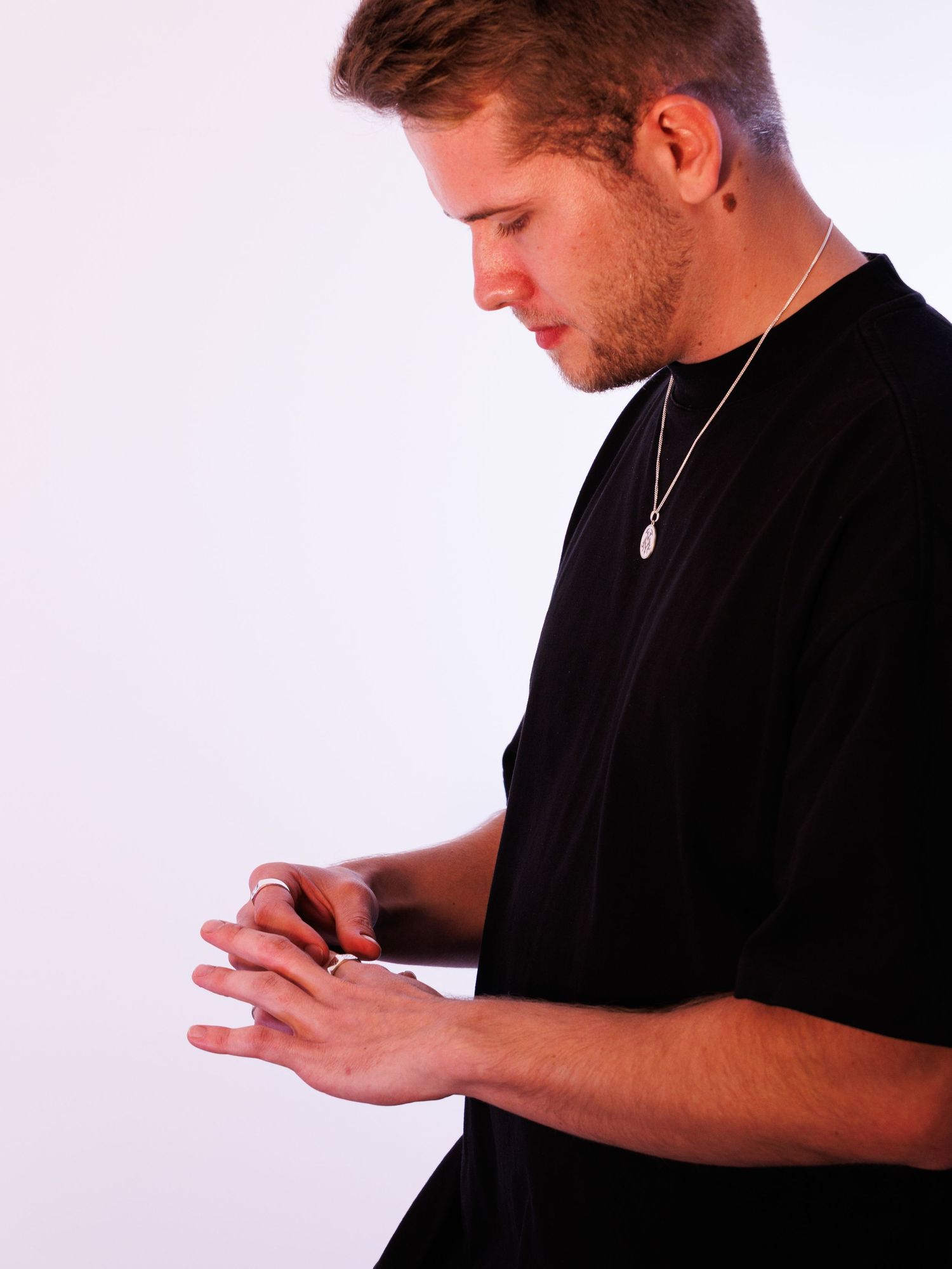 Man in black t-shirt wearing a silver pendant necklace, looking down at his hands.