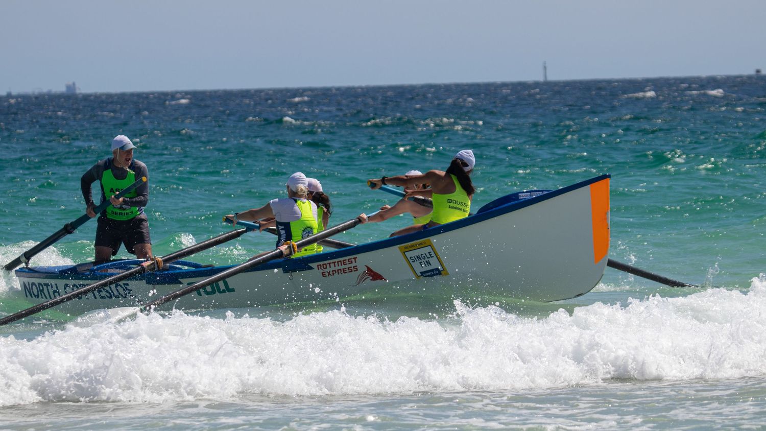 Surf lifesaving team rowing a colorful surf boat through ocean waves during a beach carnival.