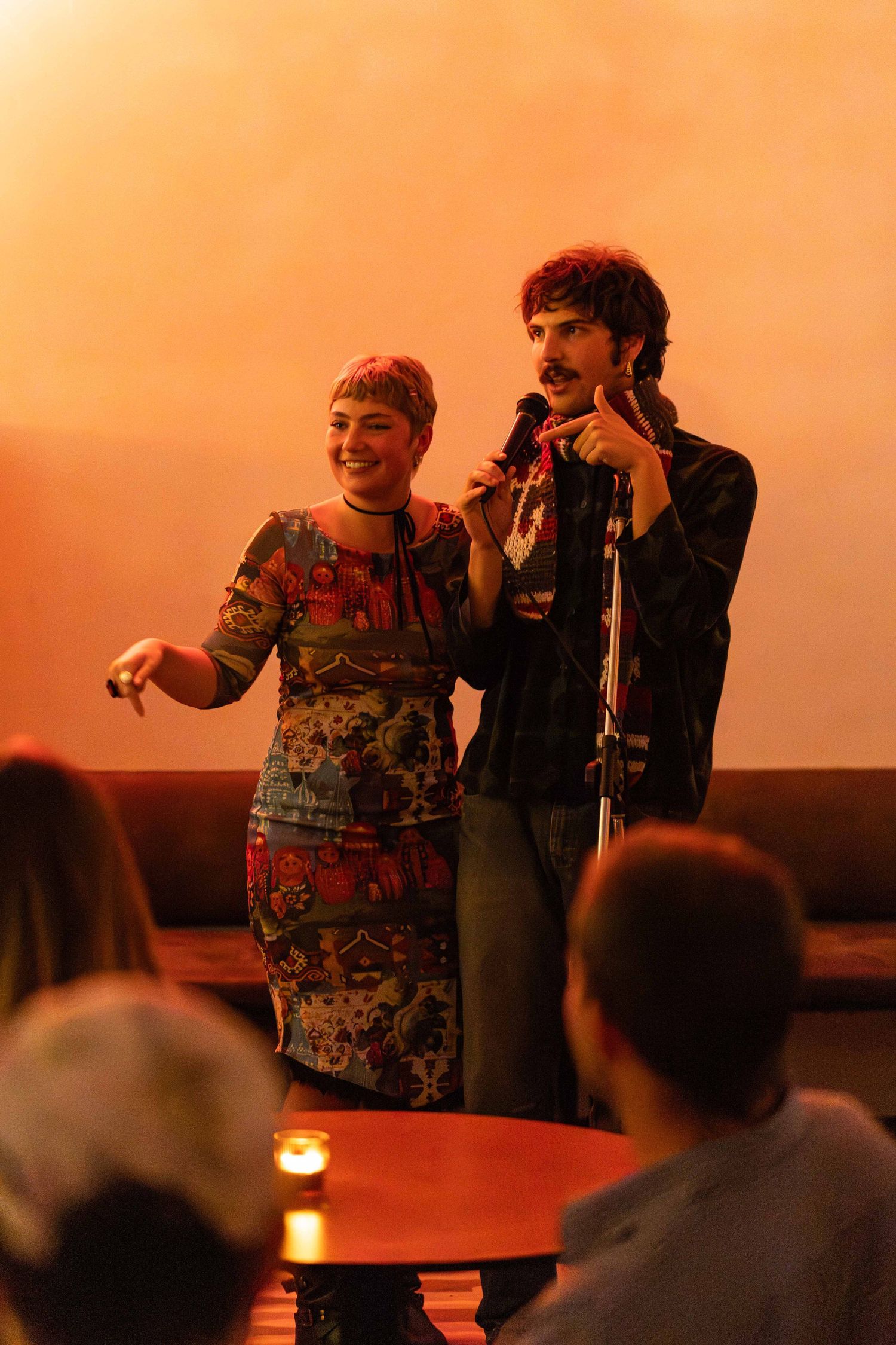 Man and woman performing together at microphone stand on warmly lit stage before seated audience.
