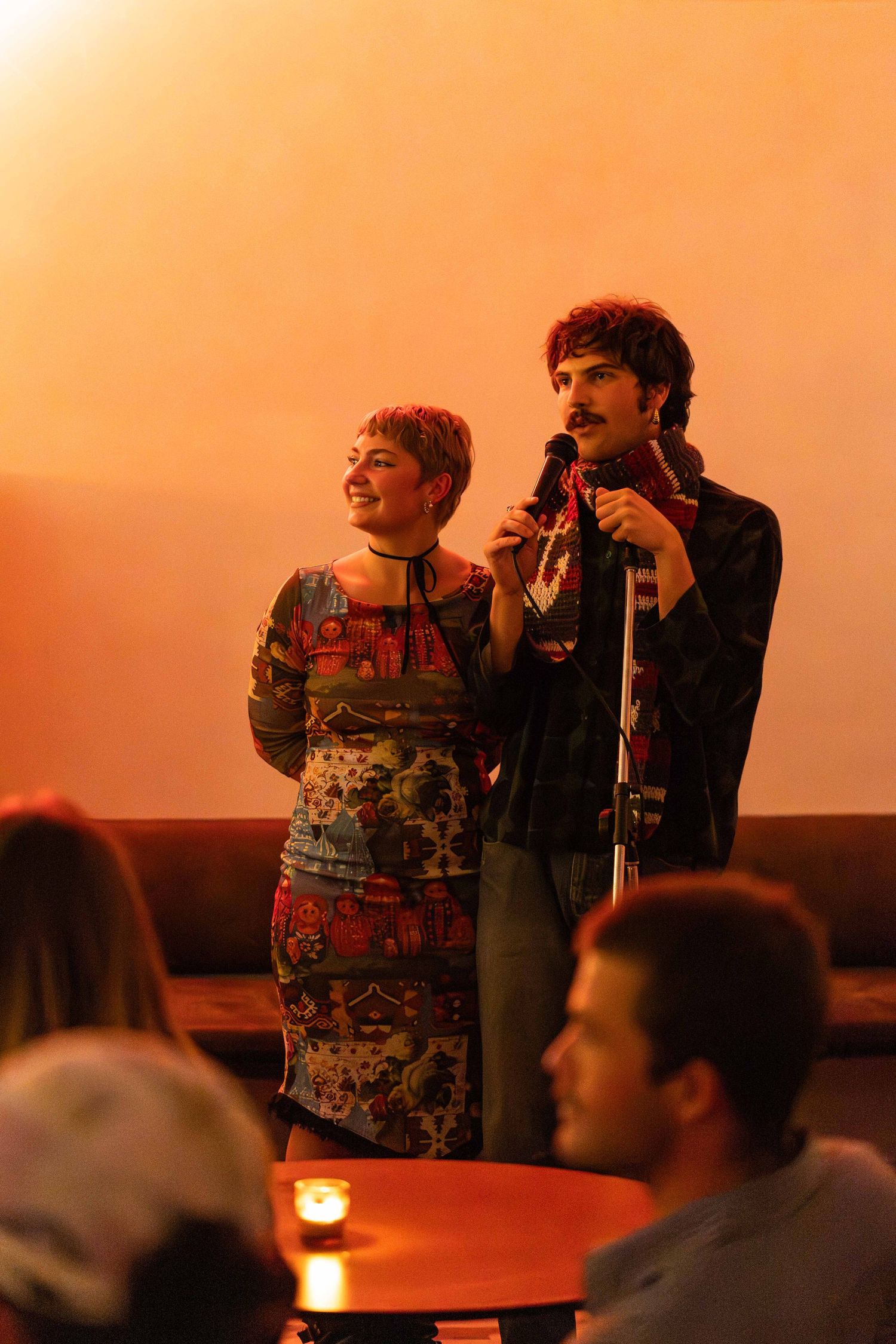A man and woman perform together at a microphone on stage, smiling in warm orange light.