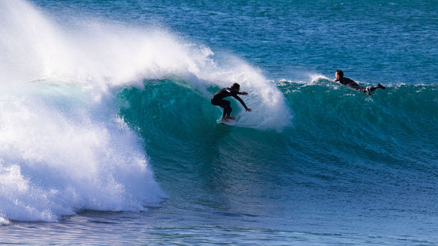 Two surfers riding a large breaking blue ocean wave with white spray on a clear sunny day.