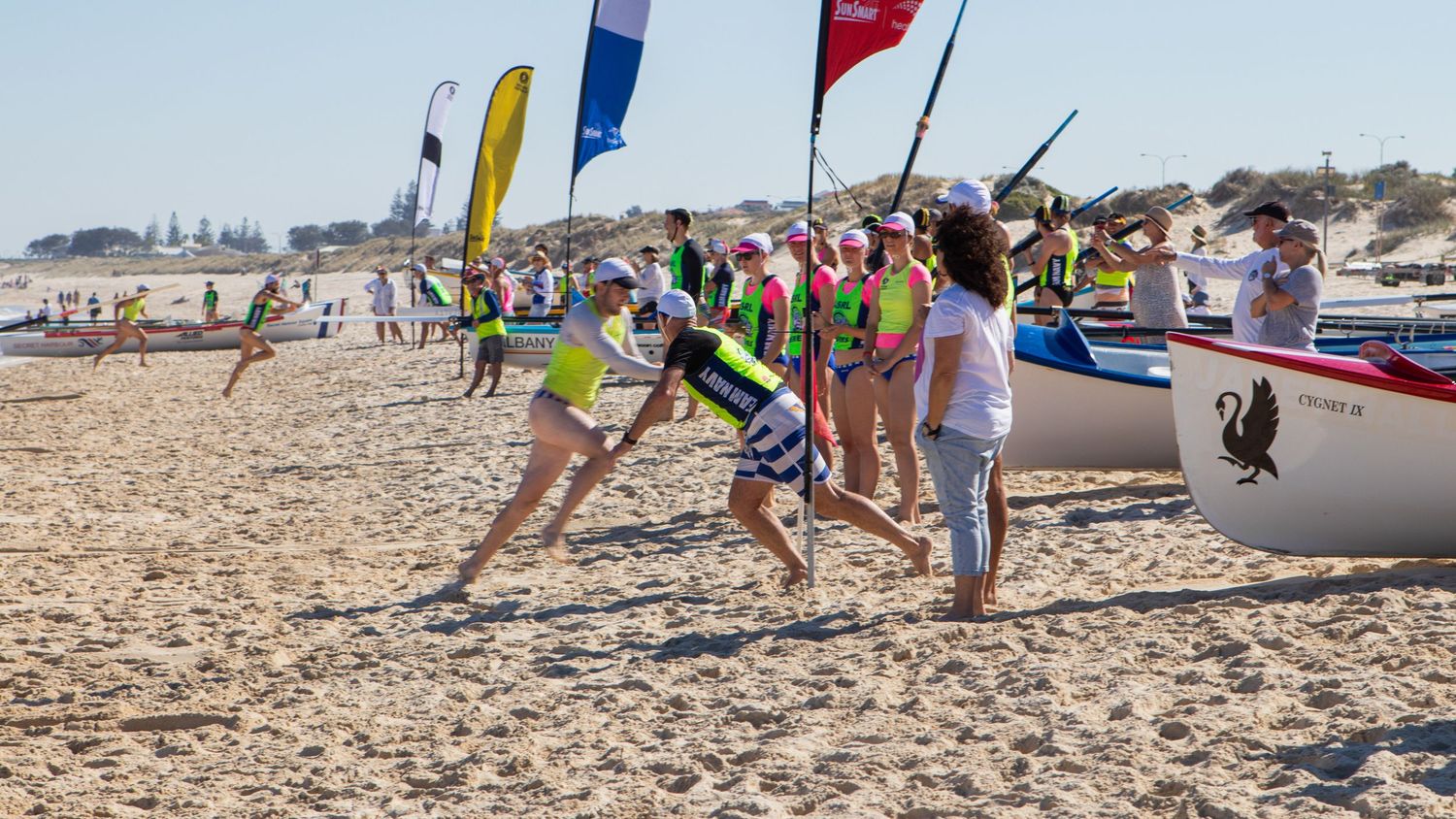 Children and adults participating in a surf lifesaving carnival on a sandy beach with colorful flags.