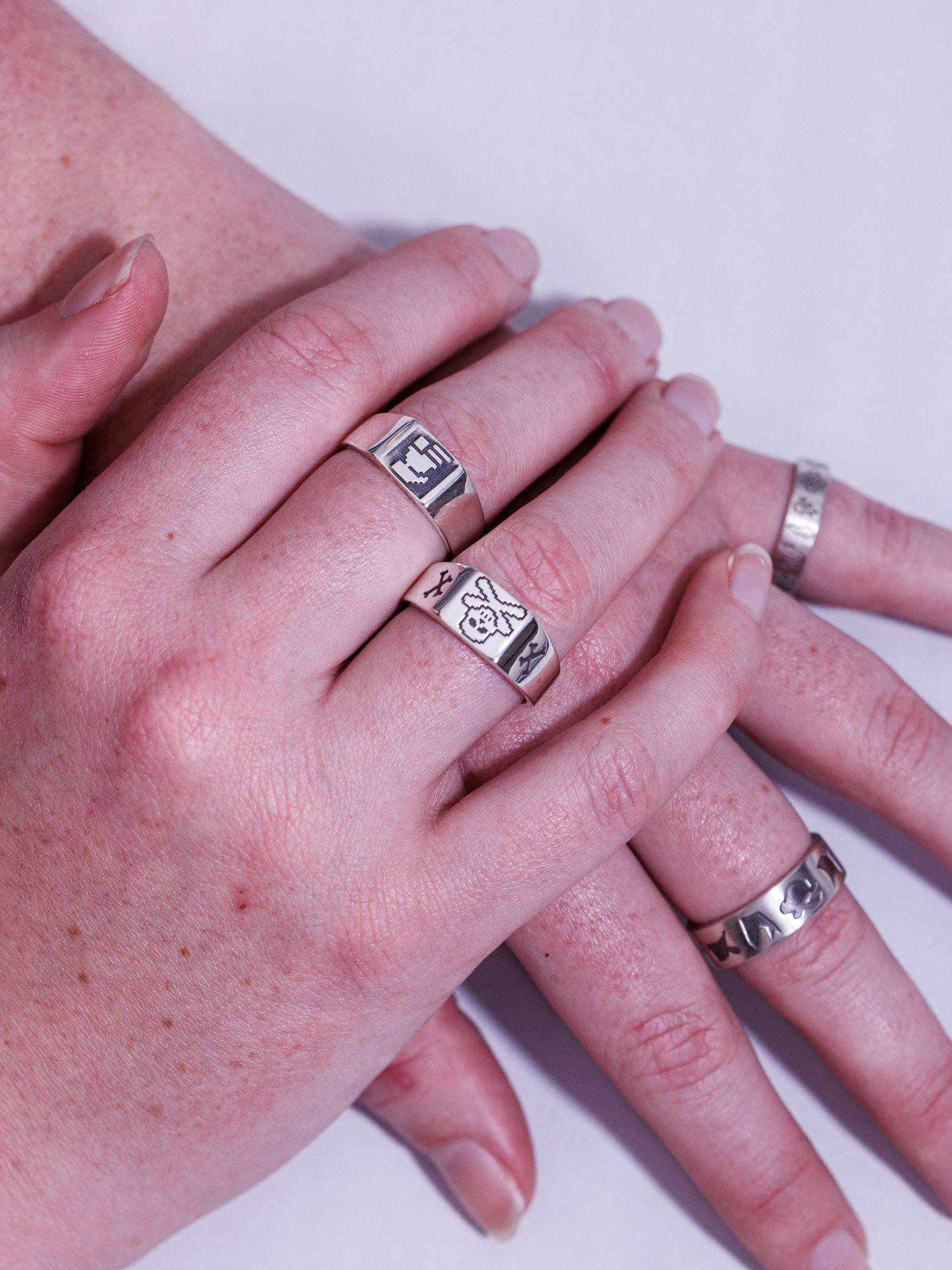 Two hands overlapping showcasing silver signet rings with engraved designs on multiple fingers.