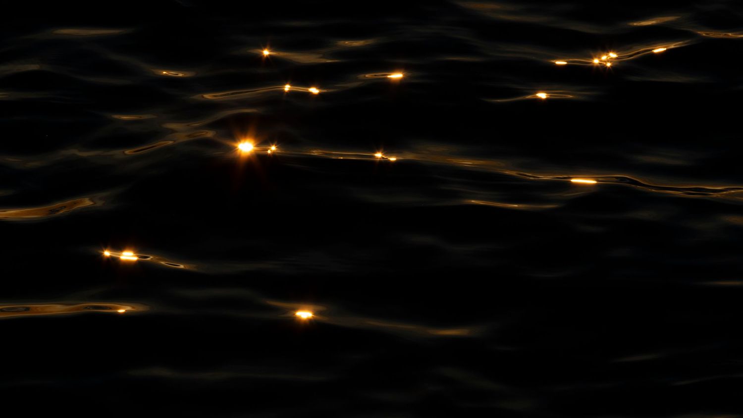 Dark ocean water at night with scattered golden light reflections glowing on gentle waves.