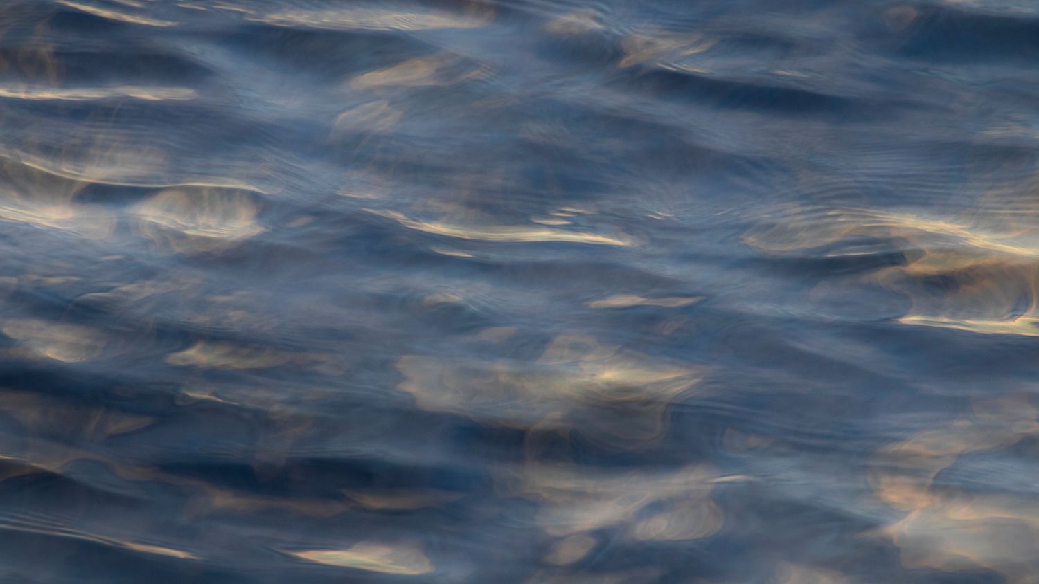Calm blue ocean water surface with soft ripples reflecting subtle rainbow hues of light.