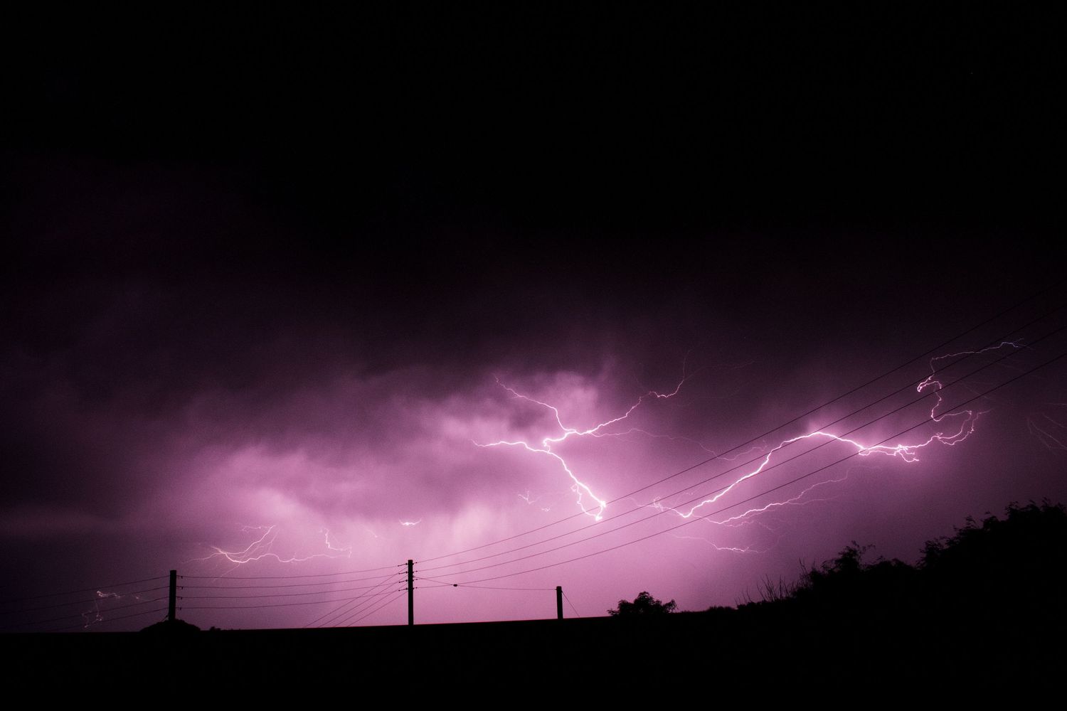Dramatic lightning storm illuminates purple night sky over silhouetted power lines and trees.