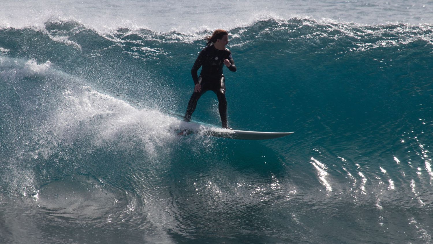 Surfer in wetsuit riding a powerful ocean wave on a white surfboard in dark blue waters.