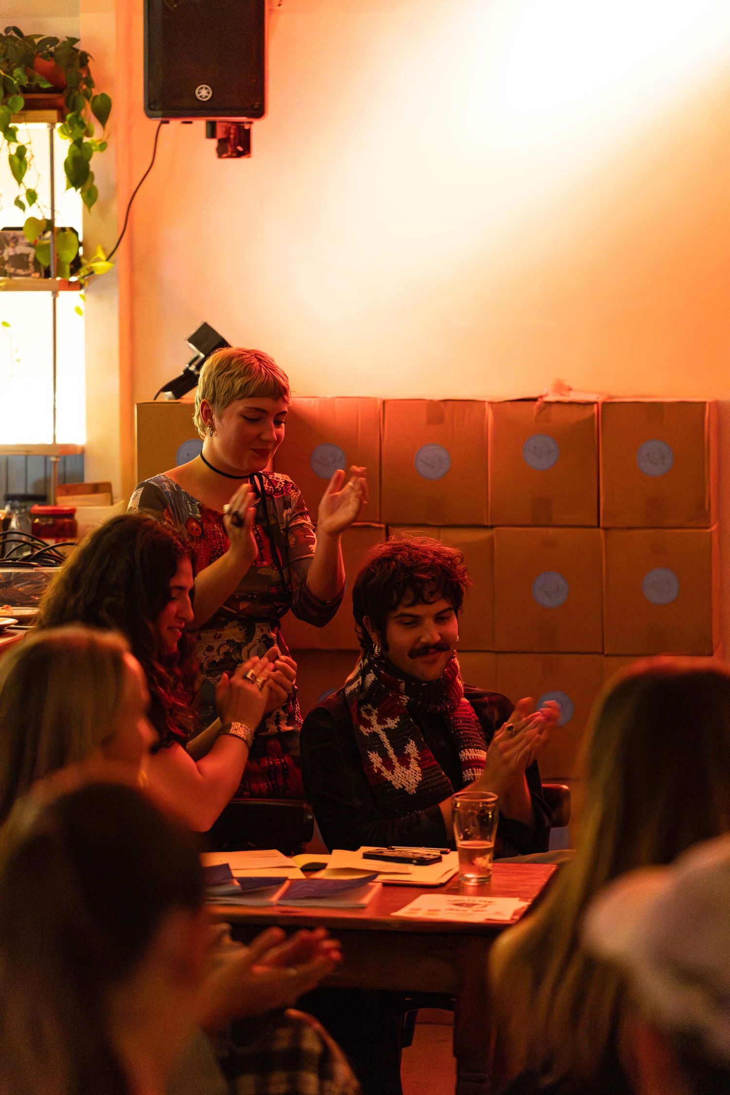 People clapping and smiling at a warm-lit indoor gathering with cardboard boxes stacked against the wall.