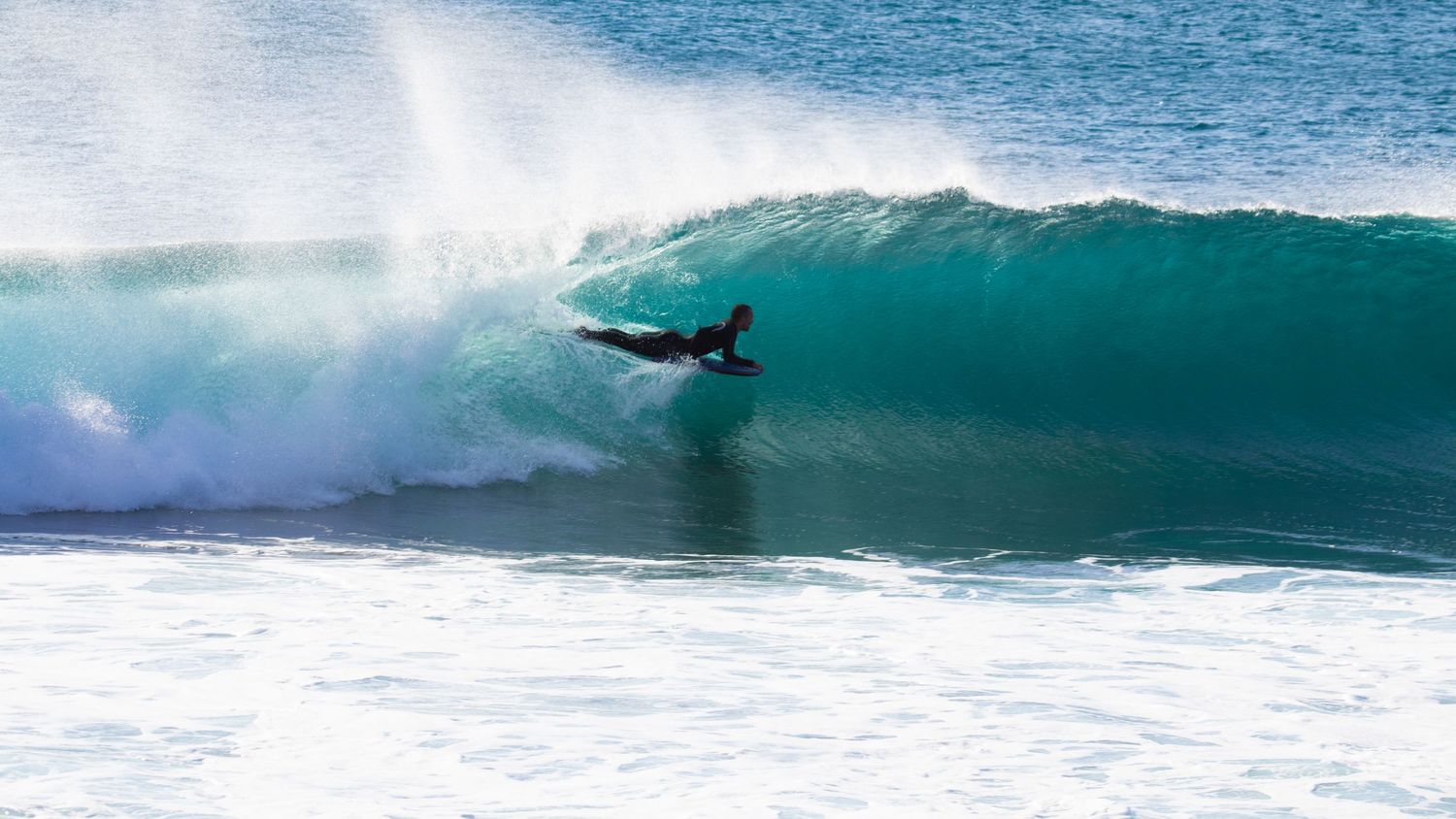 A bodyboarder rides deep inside a large turquoise barrel wave near the shore.