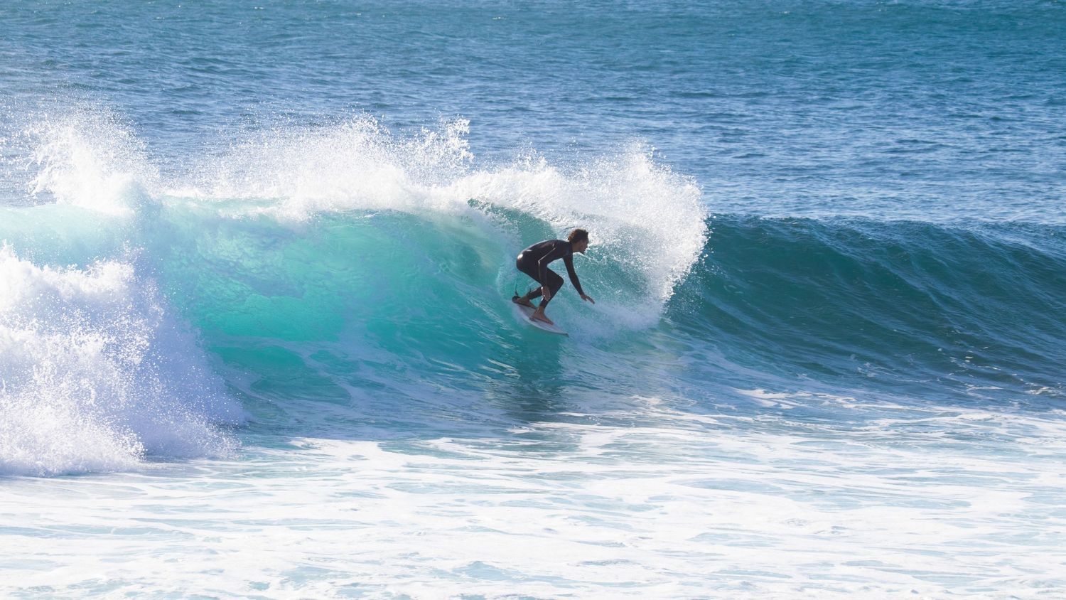 Surfer in black wetsuit riding deep inside a curling turquoise barrel wave in bright sunny conditions.
