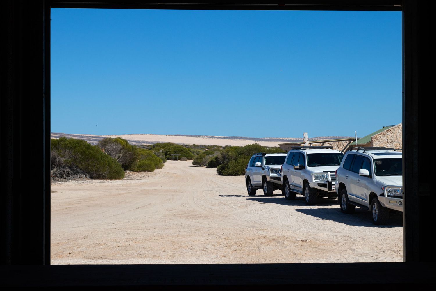 Row of white 4WD vehicles parked on remote sandy outback track viewed through dark window frame.