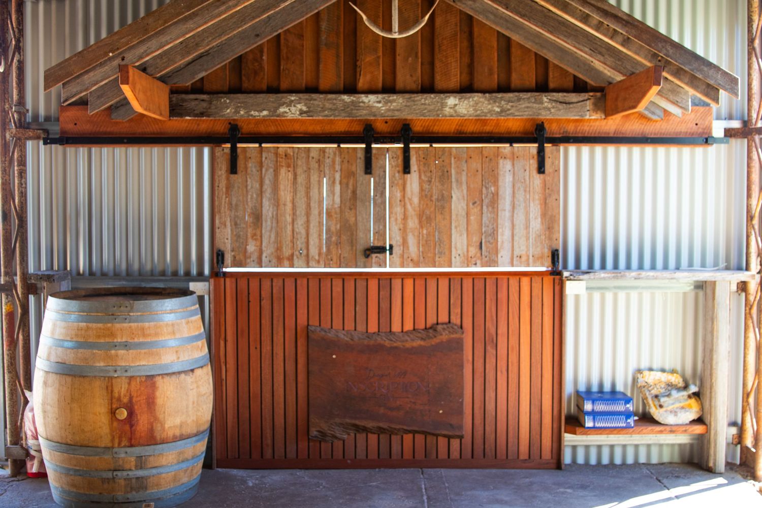 Rustic corrugated iron shed interior featuring wooden bar counter and oak wine barrel on sandy floor.