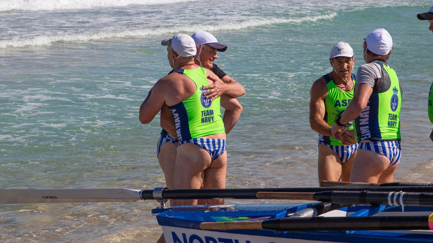 Surf boat crew members in ASRL Series bibs gather near their boat on an Australian beach.