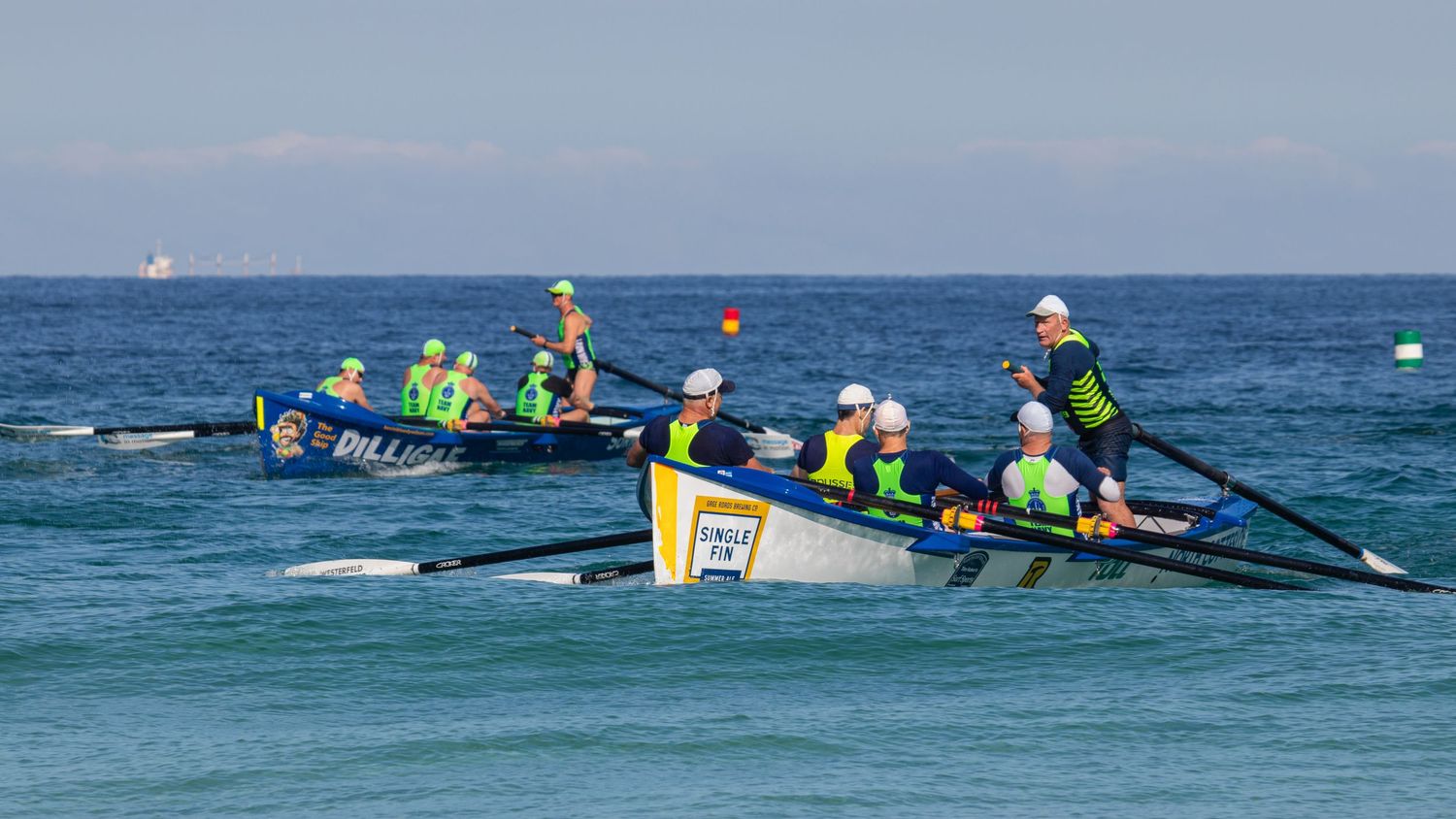 Two surf boat crews race side by side through blue ocean swells during an Australian surf carnival.