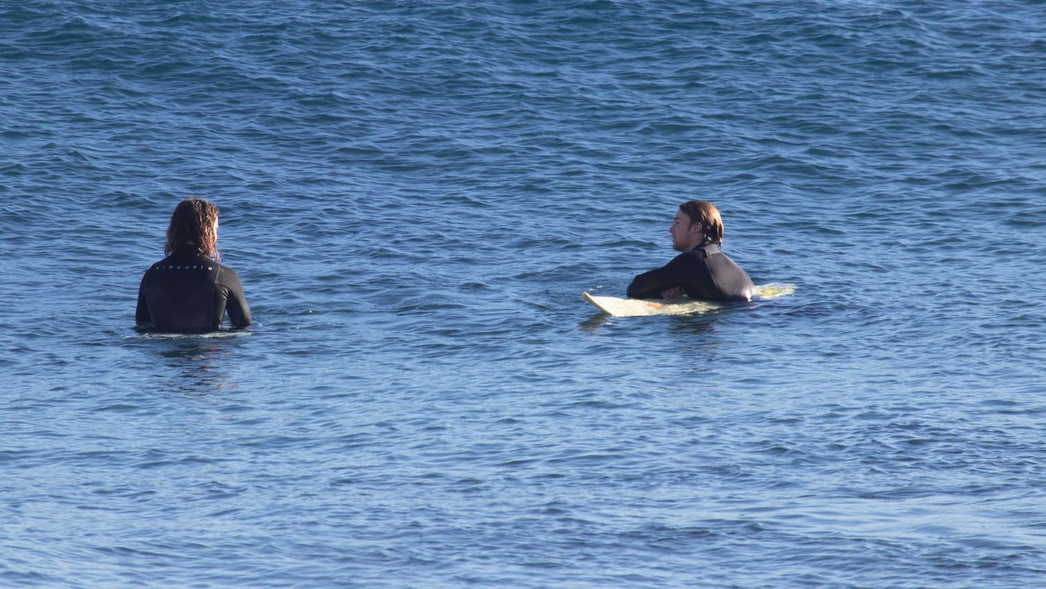 Two surfers in wetsuits sitting on surfboards in calm blue ocean water waiting for waves.