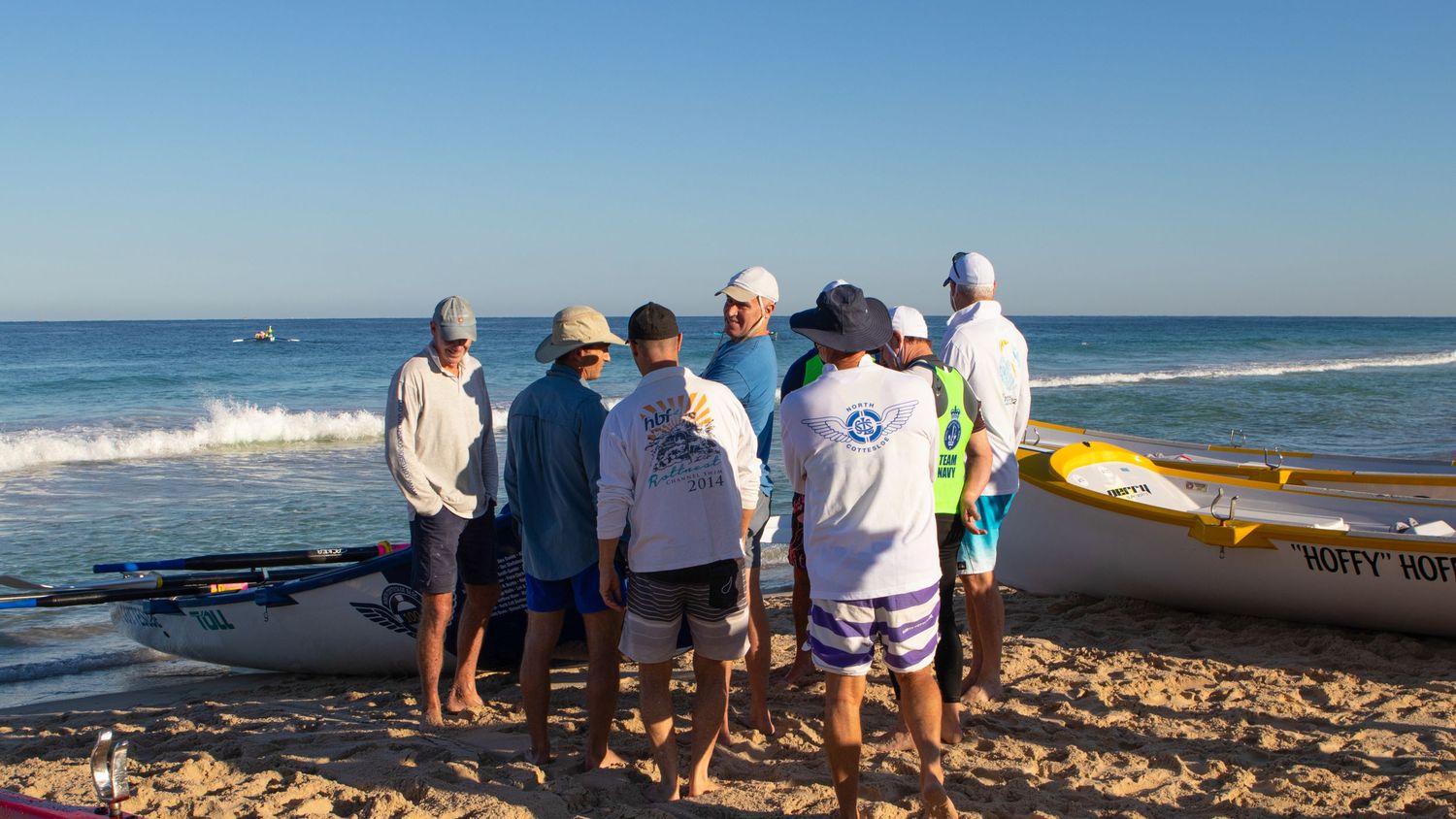 Group of surf lifesavers gathered around an outrigger canoe on a sunny beach.