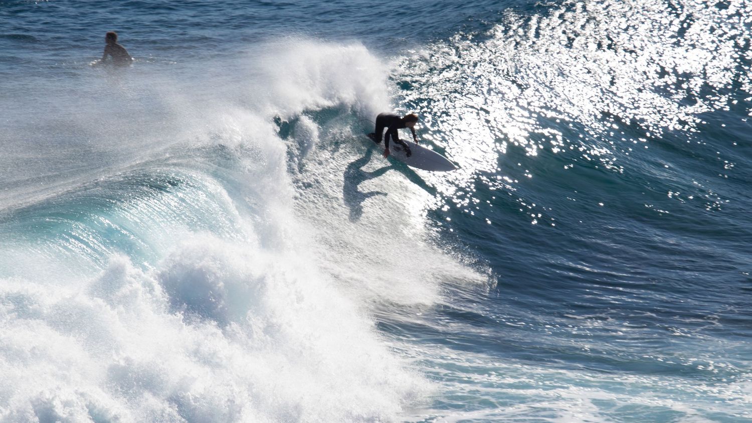 A surfer launches off a large crashing wave while another surfer watches from the water nearby.