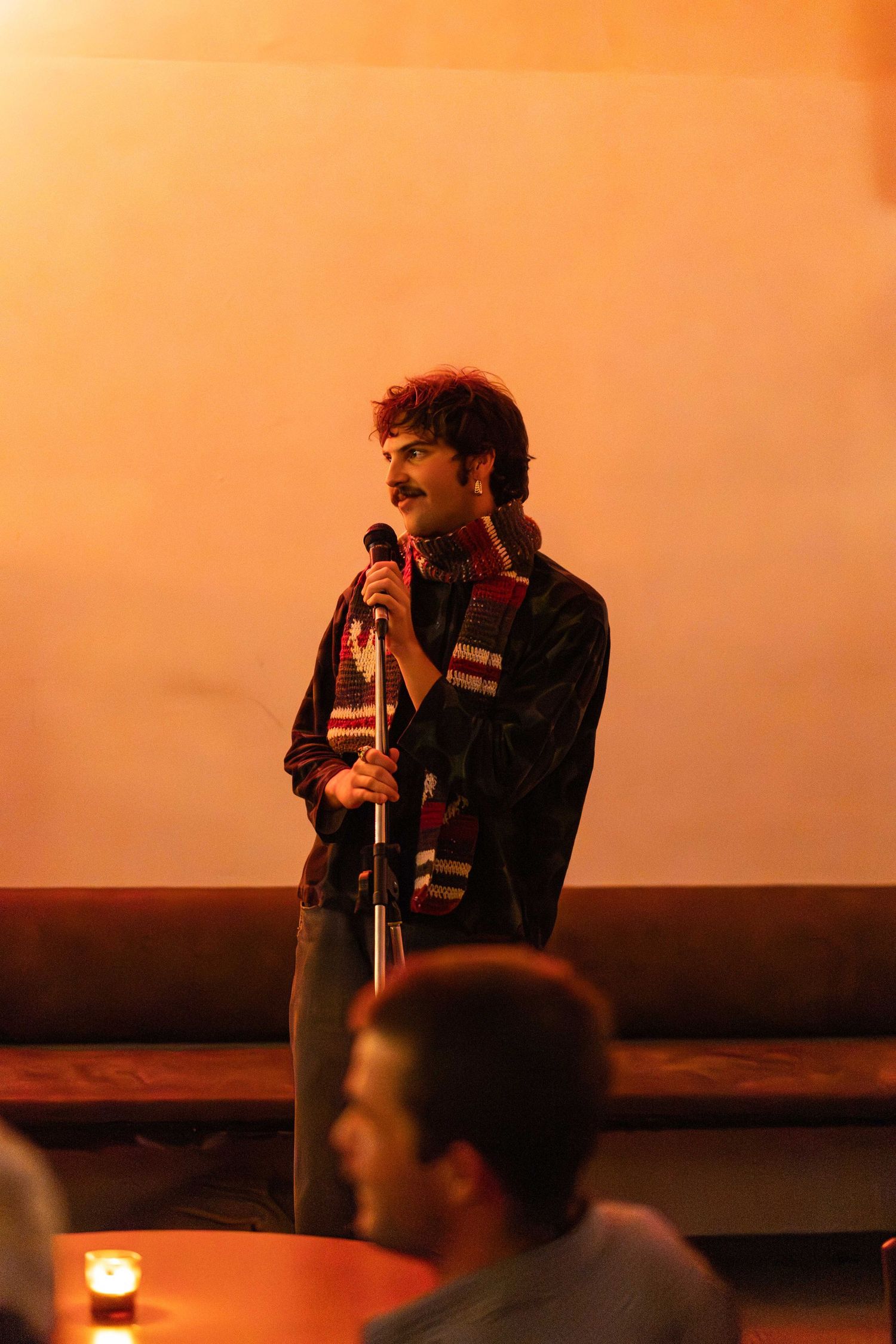 Mustachioed male performer speaking into a microphone on a warmly lit stage before an audience.