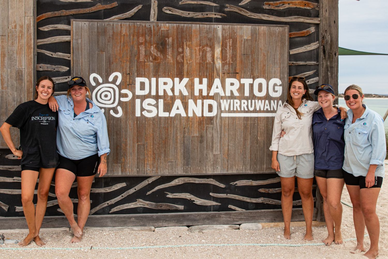 Six women posing and smiling beside the Dirk Hartog Island Wirruwana carved wooden entrance sign in WA.