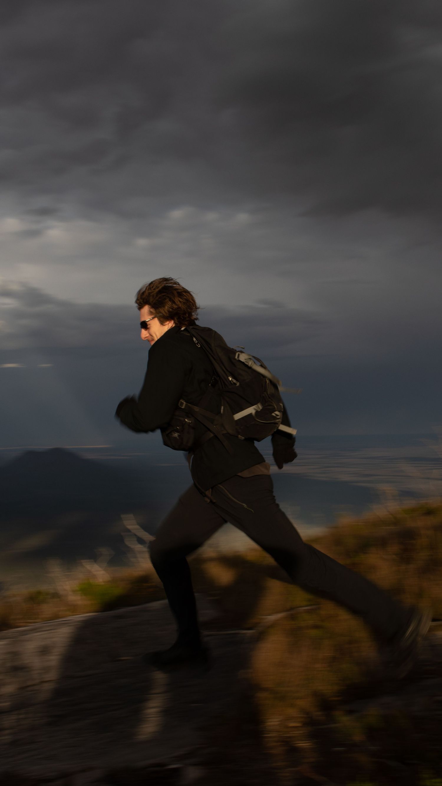 Adventurous backpacker running along mountain trail under dramatic stormy dark cloudy sky.