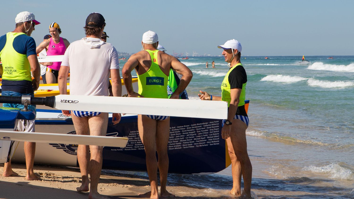 Surf lifesavers carry a rowing boat toward the ocean at a beach competition on a sunny day.
