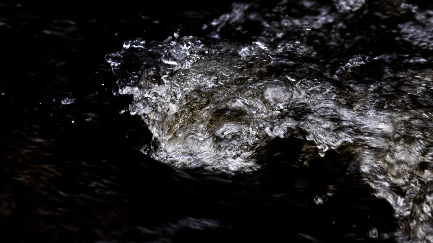 Close-up of churning white water foam swirling over dark rocks in low light.