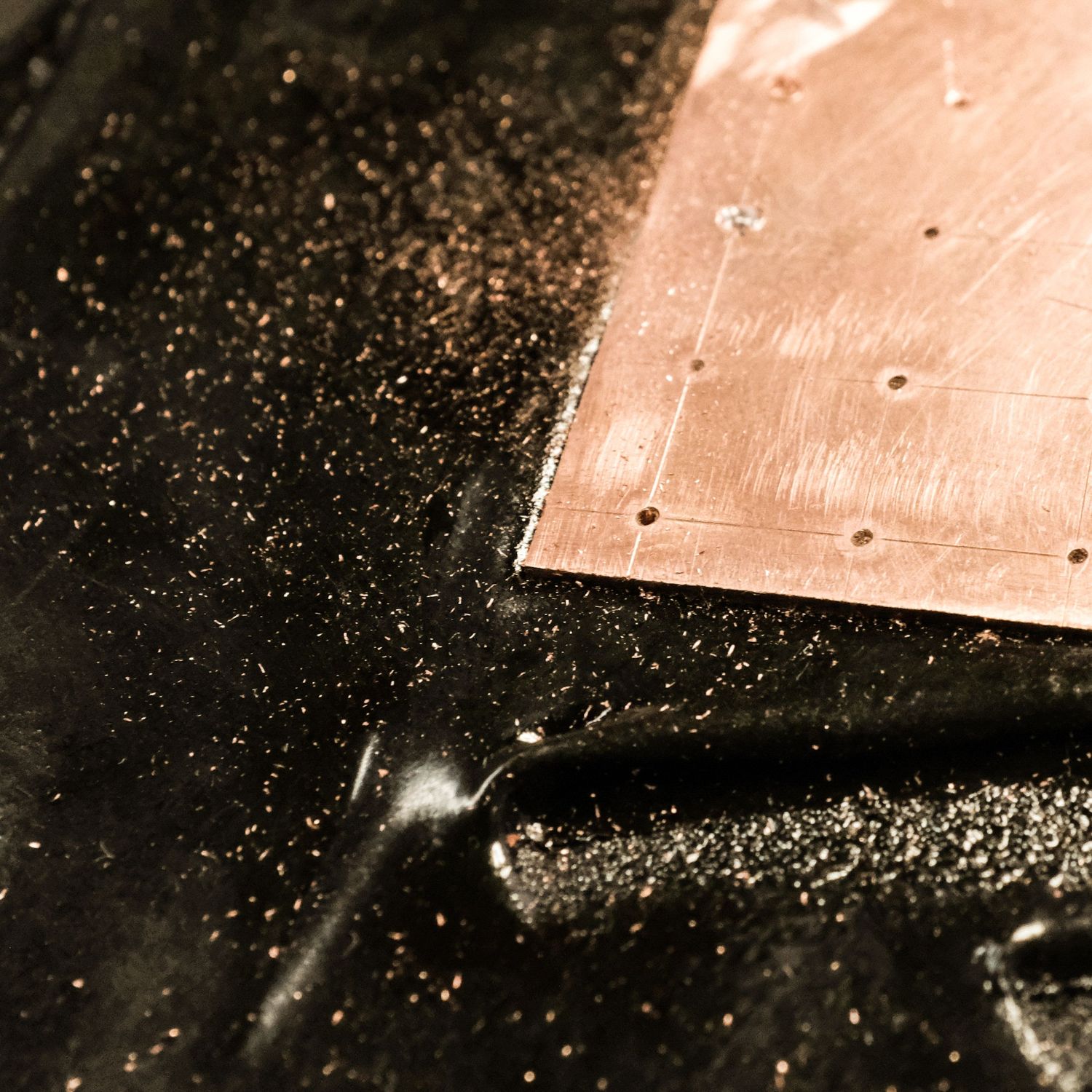 Close-up of a copper sheet with drilled holes resting on a dark, wet surface scattered with fine metallic particles.