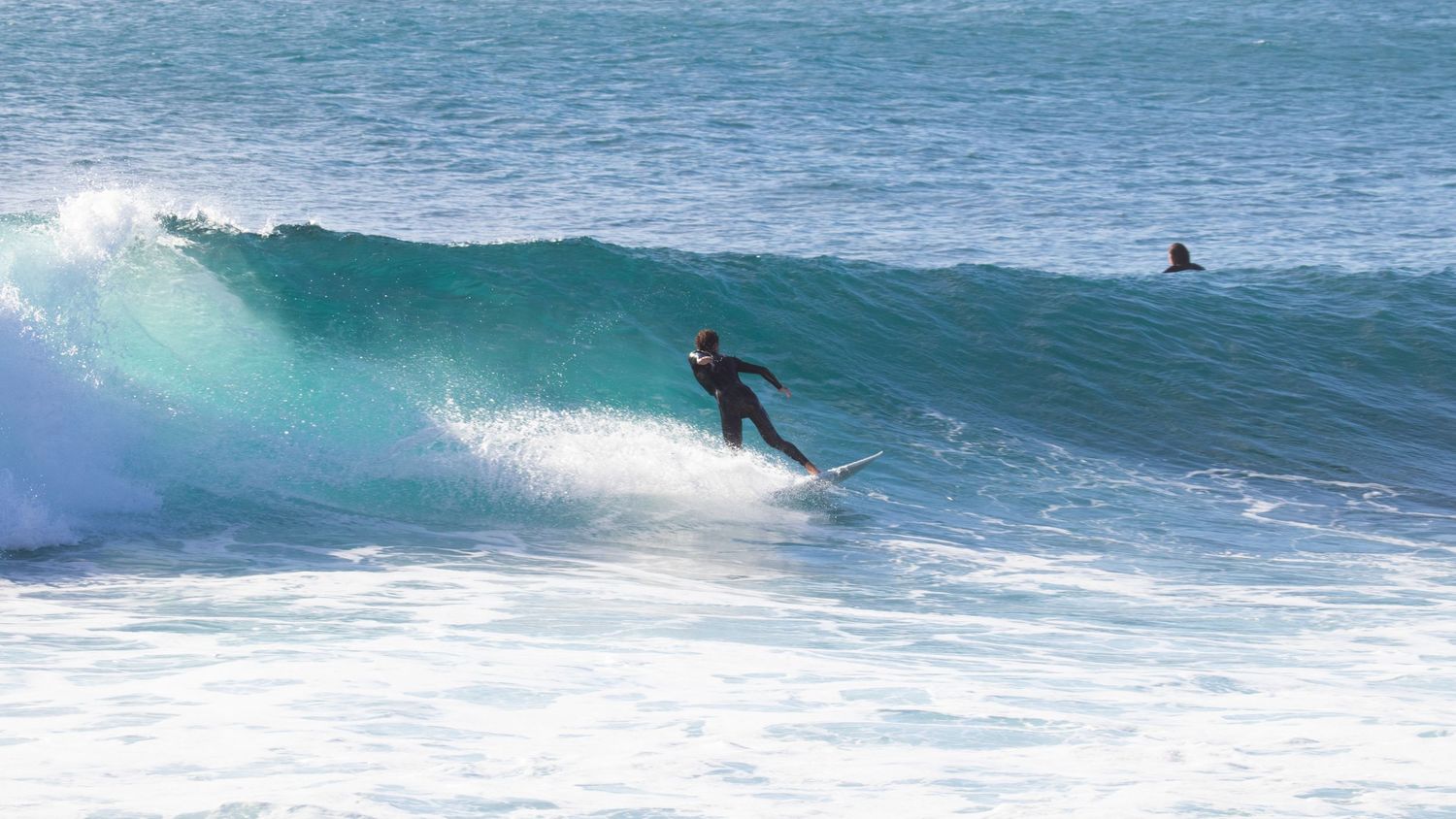 Surfer in wetsuit riding a turquoise wave with a second surfer waiting in background.