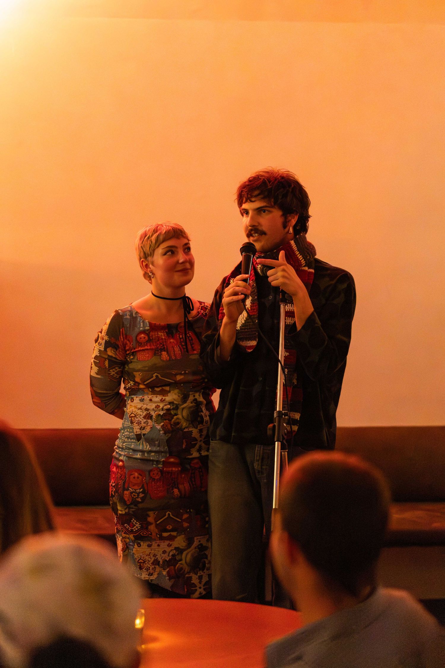 A man and woman share a microphone on a warmly lit stage during an indoor performance or spoken word event.