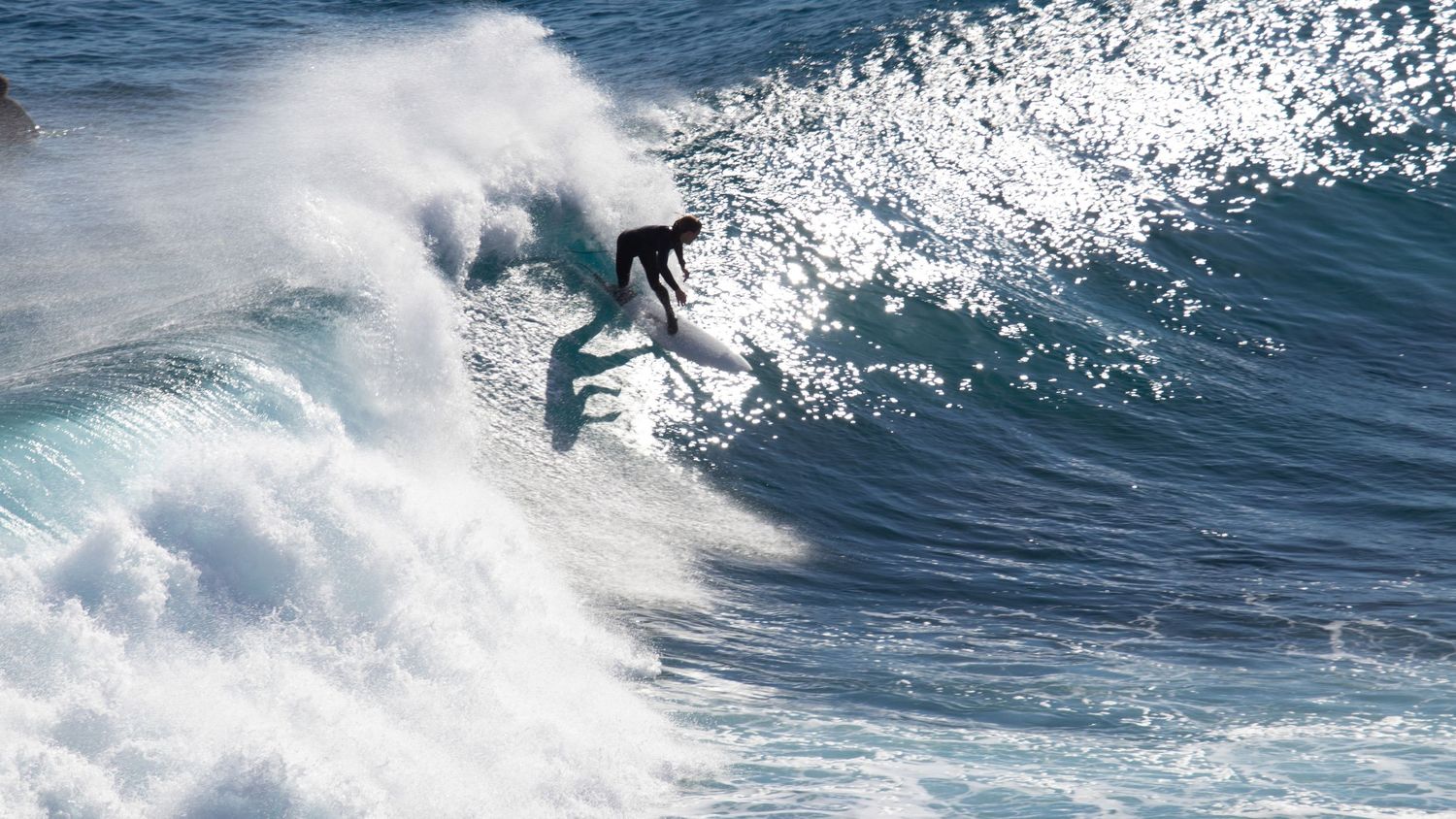 Surfer riding the crest of a large powerful white wave with dramatic ocean spray in bright sunlight.