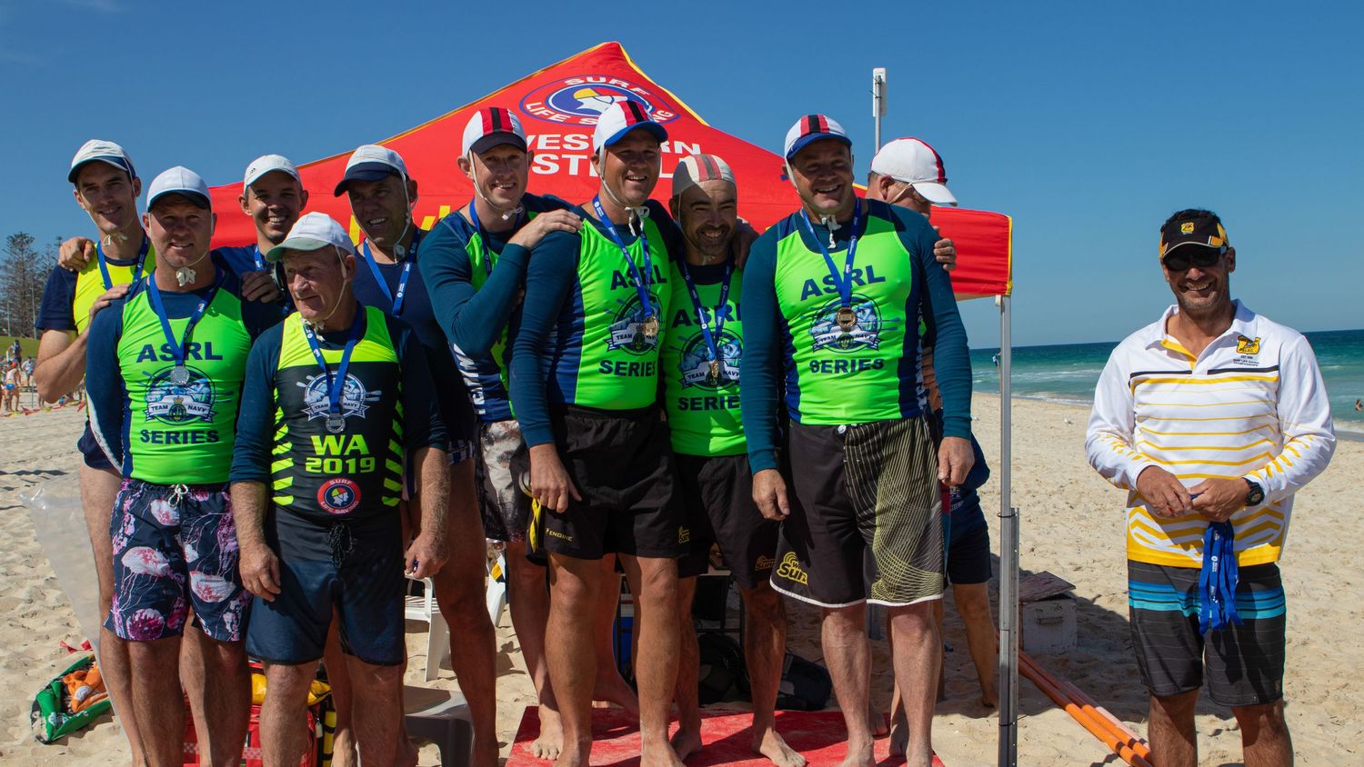 Team of surf lifesaving competitors in green uniforms posing together on beach near red event tent.