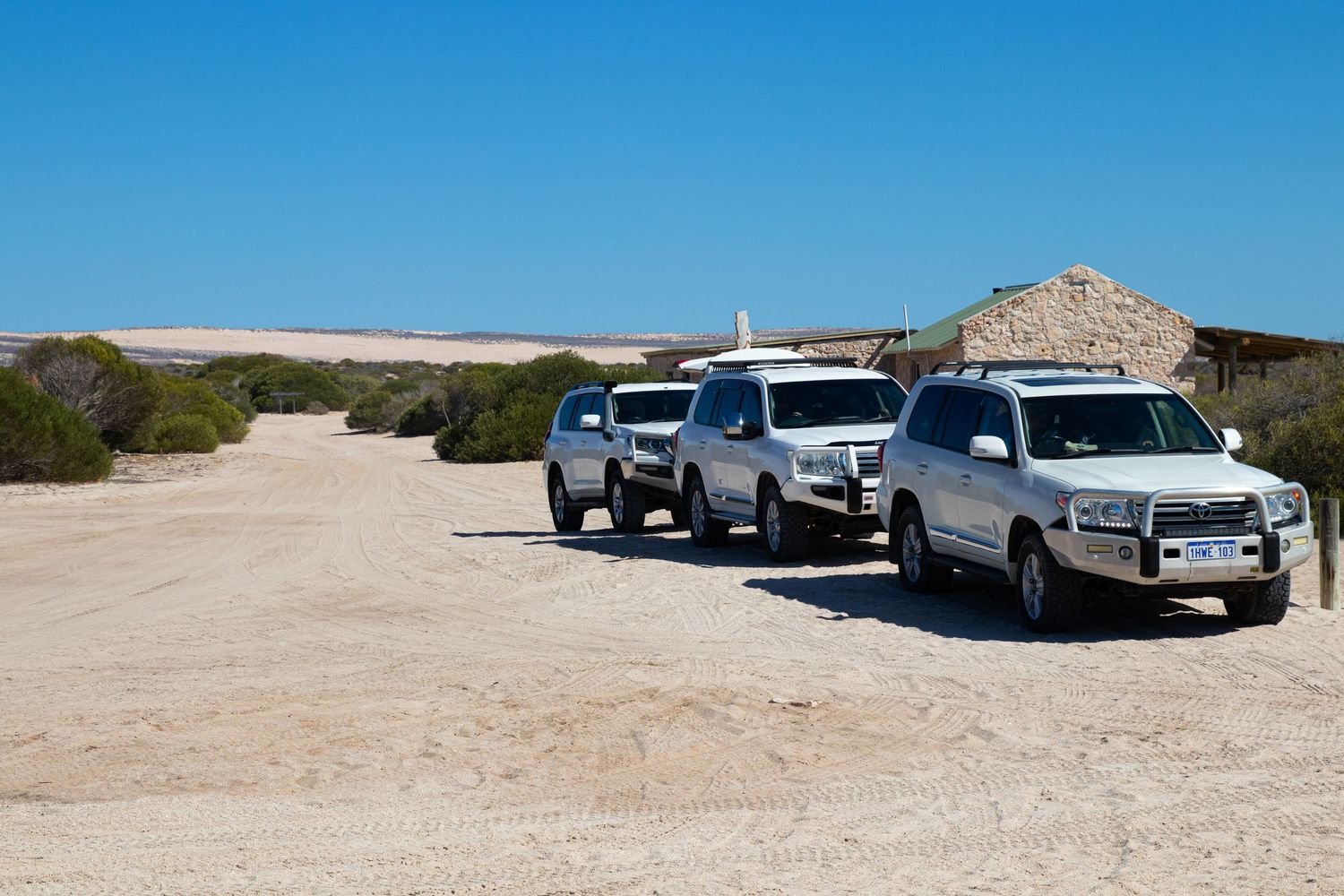 Three white Toyota Land Cruiser 4WDs parked on a sandy dirt road beside a rustic stone building.