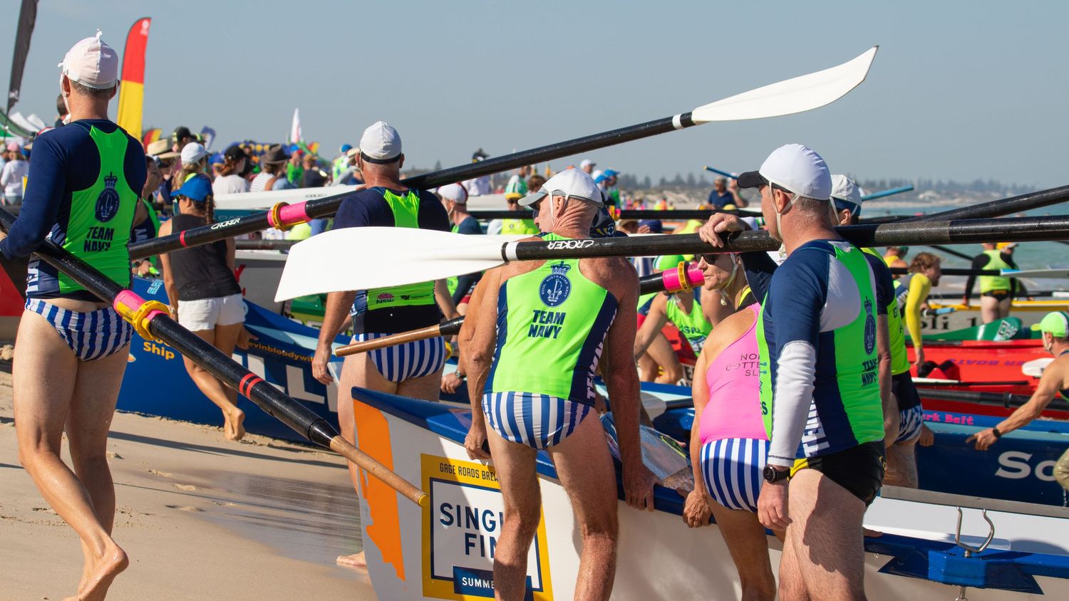 Surf lifesaving competitors in striped swimwear carry oars and boats along a busy beach carnival.